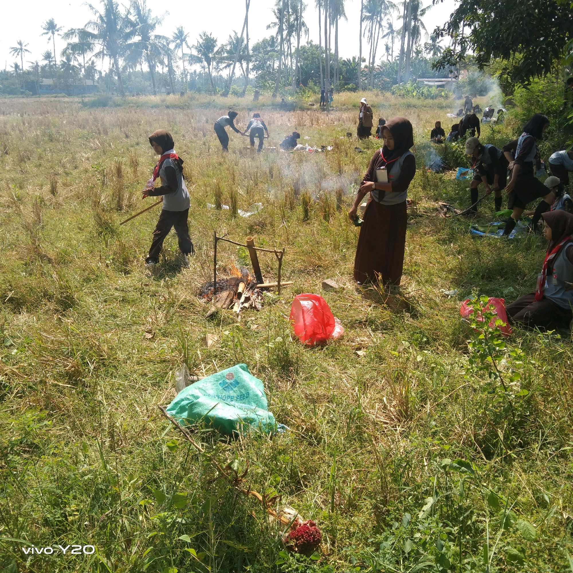 The image depicts an outdoor scene, likely in a rural or semi-rural area of Karawang, Indonesia. A group of individuals, many wearing scout-like uniforms with brown skirts or trousers and grey shirts, are gathered in a field with dry grass and some scattered wood. Some appear to be engaged in activities around a small fire pit constructed with sticks. In the background, a dense line of palm trees and other vegetation is visible, with some buildings suggesting a settlement nearby. The overall atmosphere appears to be a daytime activity or event. There are also several plastic bags, including a green one and a red one, present on the ground, indicating some level of human presence and activity.