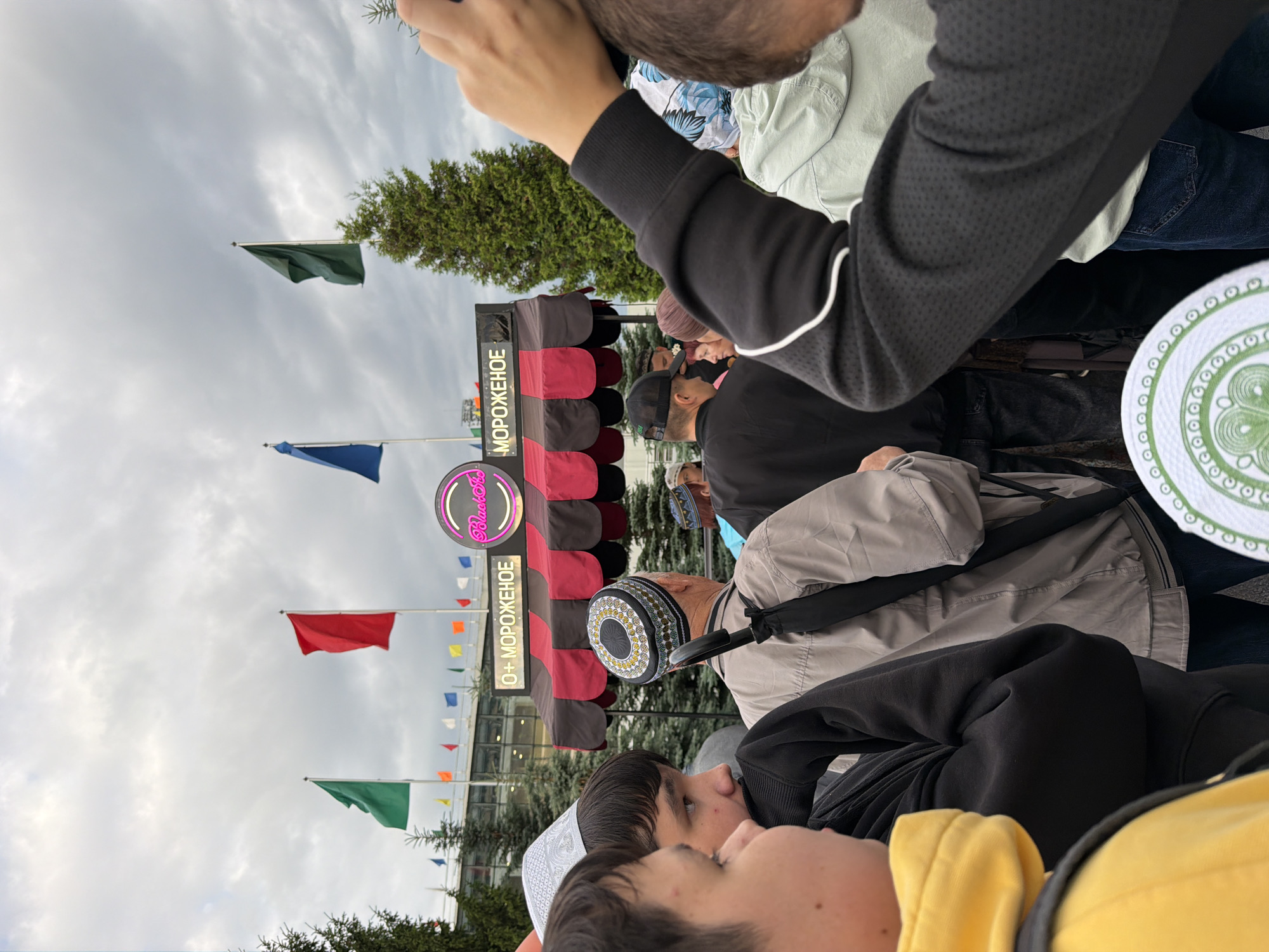 In an outdoor urban setting, a crowd of people, some wearing traditional headwear, gathers in front of an awning that reads "O+ МОРОЖЕНОЕ" and features a circular pink neon sign with the word "BlackSea". Several flags, including red, green, and blue, are visible in the background against a cloudy sky, flanking tall evergreen trees. The people in the foreground are casually dressed, with some in hoodies and jackets. The scene appears to be a busy outdoor event or market, possibly a food stall or festival.