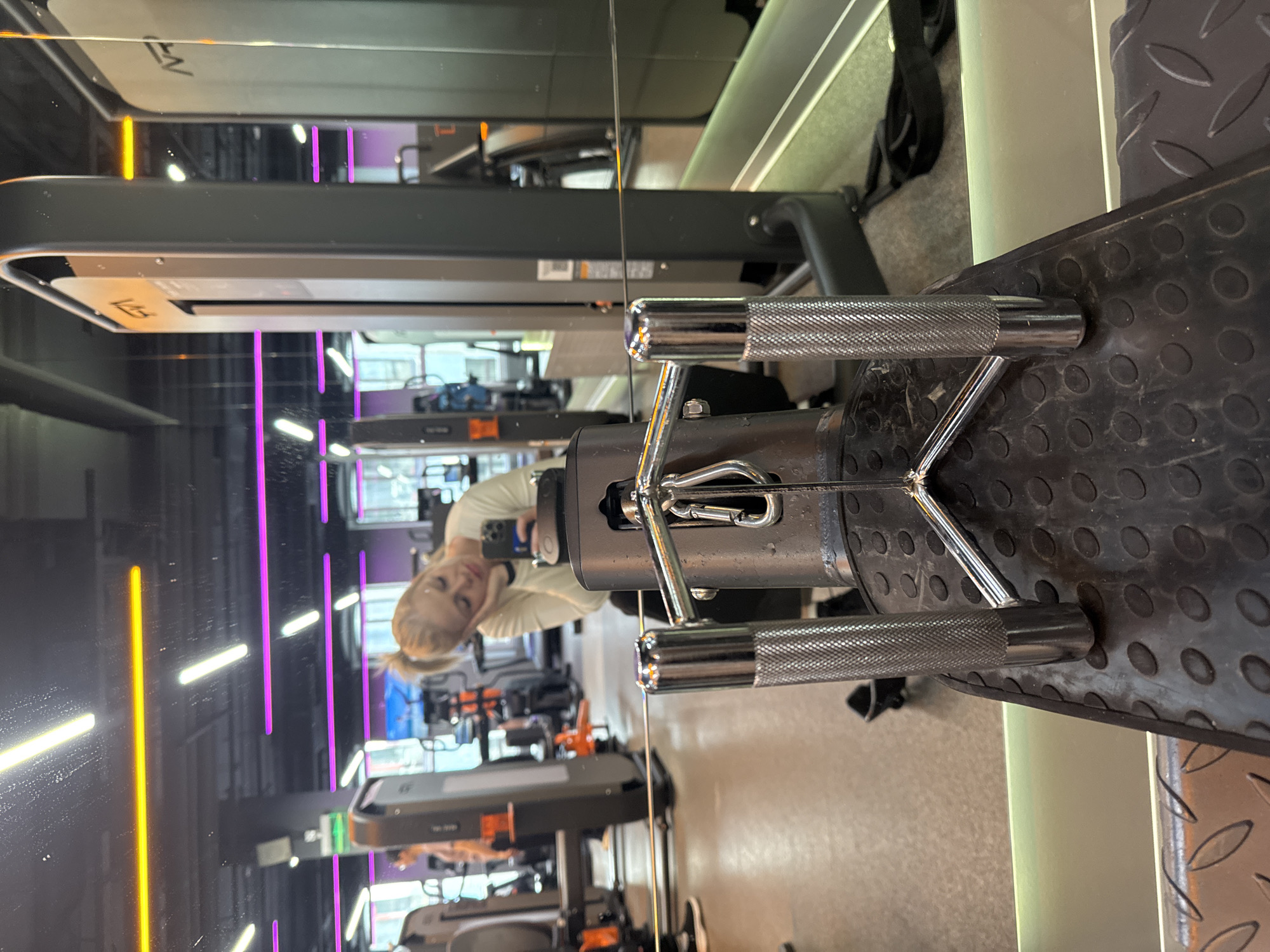This indoor shot in what appears to be a gym in Saint Petersburg, Russia, shows a chrome-handled exercise machine centered in the frame. The machine is situated on a black textured platform, and its chrome bars are prominently displayed. A person, reflected in a mirror, is visible in the background, holding a phone and looking at its screen. The gym features a modern interior design with overhead lighting and other exercise equipment arranged in rows, also visible in the reflection. The lighting in the gym includes neon-colored accents.