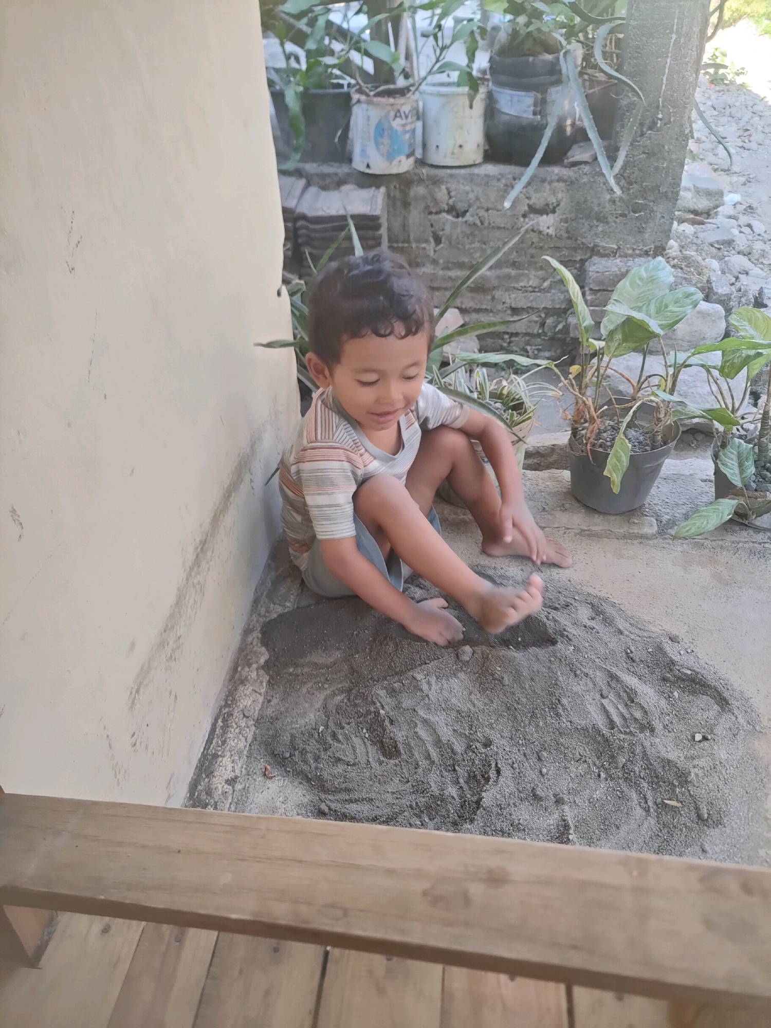 A young boy is playing in a pile of sand next to a concrete wall. He is wearing a striped t-shirt and shorts, and he is sitting on the ground with his legs crossed. The sand is gray and appears to be mixed with small stones. In the background, there are several potted plants, some of which are in plastic containers. A wooden railing runs along the bottom of the frame. The scene appears to be outdoors, possibly in a residential area in Nganjuk, Indonesia, given the dry, dusty environment and the style of construction.