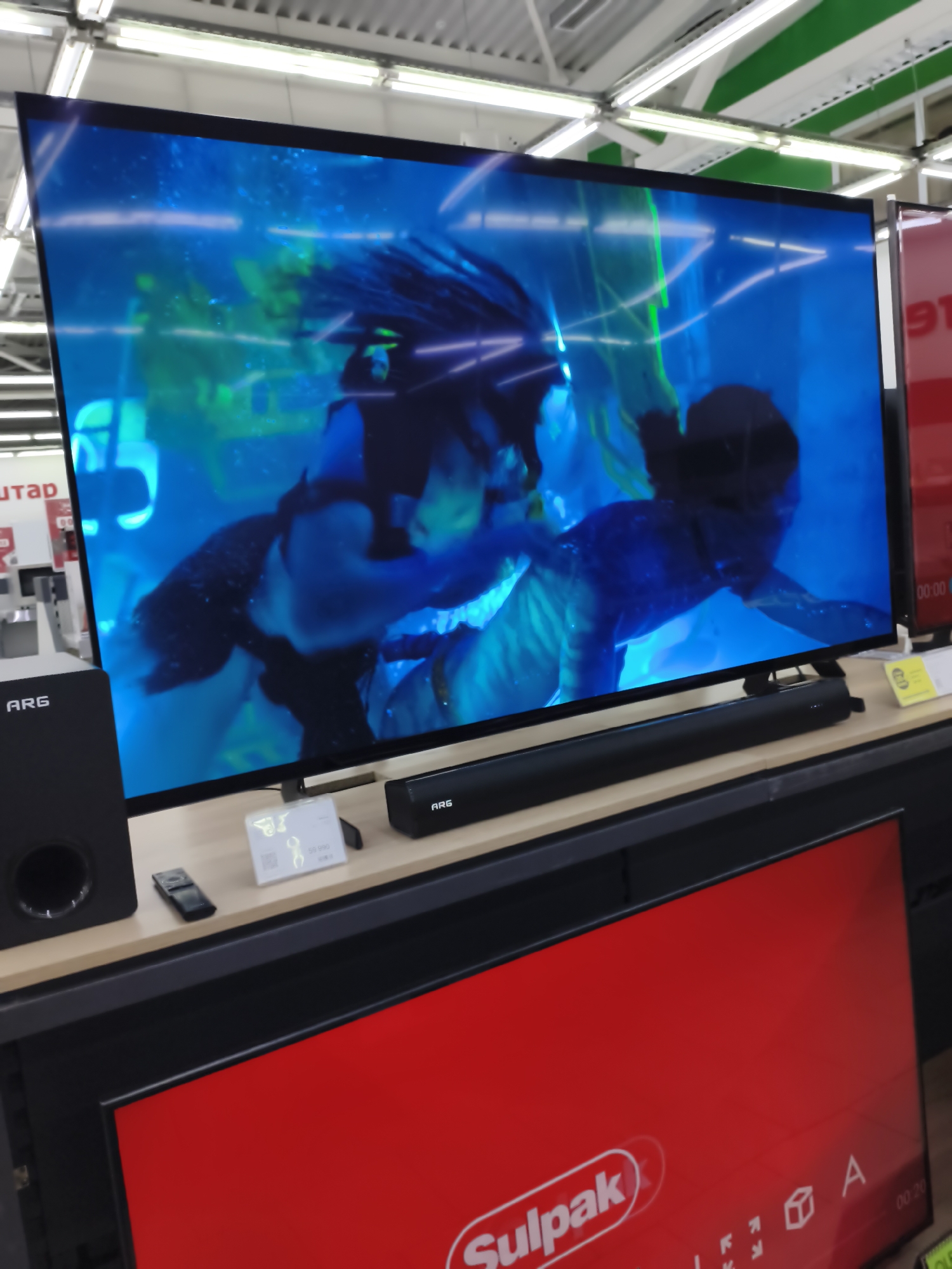 A wide-angle, slightly angled shot captures a large, flat-screen television displaying a vibrant blue scene with two figures, possibly underwater, on a display stand in what appears to be a retail electronics store. The television is the dominant subject, angled towards the viewer. To the left of the television is a black subwoofer with the "ARG" logo visible. In front of the television is a black soundbar, also with the "ARG" logo. A small white price tag with what appears to be a QR code and pricing information is placed in front of the television. To the right of the soundbar, a red sign is partially visible. In the foreground, another large red display screen is visible, featuring the "Sulpak" logo. The background shows the interior of a store with bright fluorescent lighting and some shelving units. The scene suggests a retail environment, possibly a Sulpak electronics store in Aktobe, Kazakhstan, given the prominent Sulpak branding on the foreground display. The overall lighting is bright and even, indicative of an indoor commercial setting.