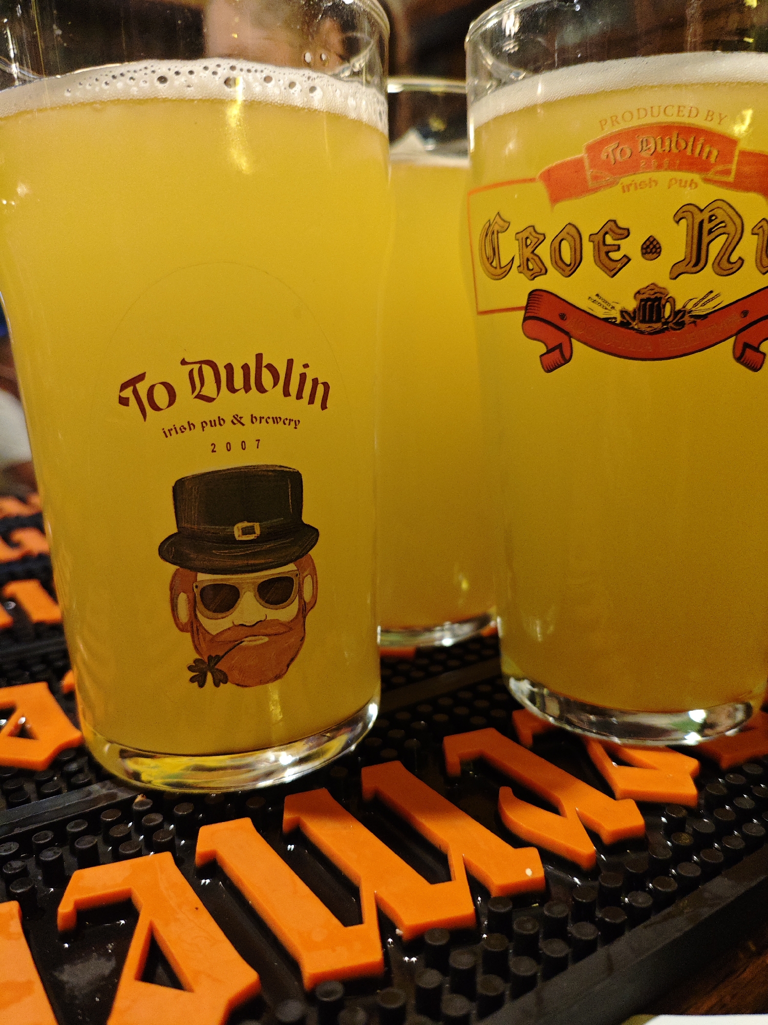 The image depicts three glasses of yellow, frothy beverage, likely beer, placed on a black bar mat. The glasses are decorated with labels. The leftmost glass has a label with the text "To Dublin irish pub & brewery 2007" and a cartoon drawing of a leprechaun with a hat, sunglasses, and a clover stem in his mouth. The rightmost glass has a label that reads "PRODUCED BY To Dublin irish pub" at the top, and "Croé N" in large decorative lettering with a red ribbon below it. The background is blurry, showing hints of a person and possibly a bar environment. The orange letters "MAN" are visible on the bar mat in the foreground, partially obscured by the glasses. The image suggests a setting in a pub or brewery, potentially related to Irish culture given the labels. There is no specific information to determine the city of Kyiv, Ukraine from the image itself.