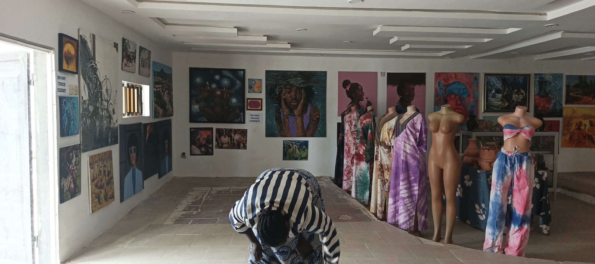 The image displays the interior of an art exhibition or gallery space, consistent with a cultural setting in Akwanga, Nigeria. The bright, white walls are densely covered with a diverse collection of paintings of various sizes and styles. Many artworks feature portraits of Black individuals, often with vibrant colors and expressive themes, including a large painting of a person partially submerged in water. Other paintings depict cultural scenes, abstract forms, and landscapes.
In the foreground, a person, likely female, with braided dark hair, is bent forward, facing away from the viewer. They are dressed in a blue and white garment, featuring stripes on the upper portion and a patterned design on the lower half, characteristic of traditional African attire. Their exact activity is unclear, but they appear to be inspecting or adjusting something on the tiled floor.
The right side of the room showcases fashion and pottery. Several mannequins display colorful tie-dye or batik garments, including long, flowing dresses in shades of pink, green, yellow, and purple. Two bare-chested female mannequins are also present; one stands near the dresses, while another wears a vibrant pink and blue tie-dye two-piece outfit (bandeau top and loose trousers) next to a display table. This table holds various terracotta pots and ceramic items.
Natural light floods the room from a large, tall window on the left, indicating it is daytime. The ceiling features modern architectural details with recessed lighting and multi-layered rectangular panels. A small white sign affixed to the left wall clearly states, "DO NOT TOUCH EXHIBIT." The floor is tiled in a light color, with some areas in the center showing signs of wear.