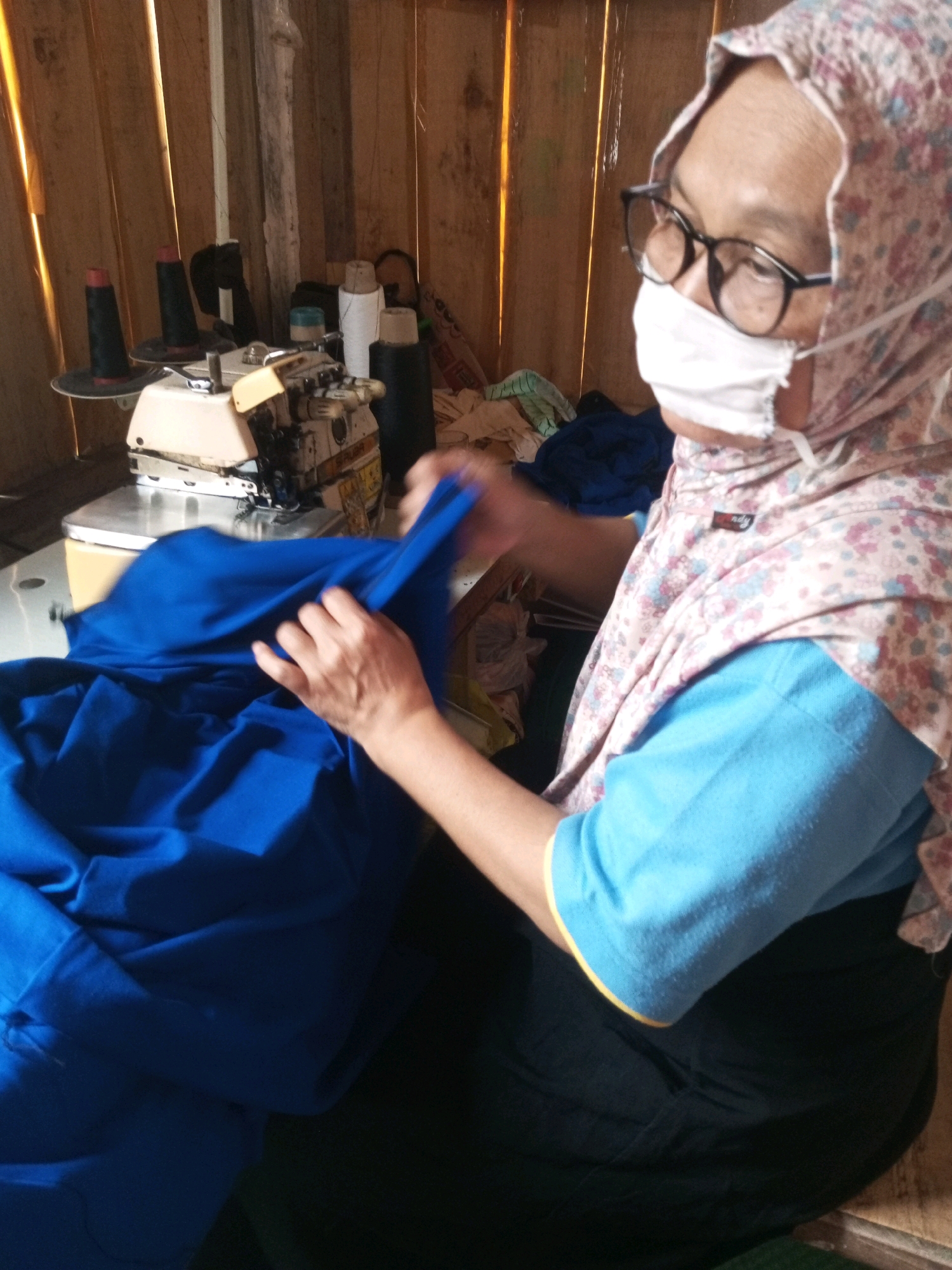 In Kalibeber, Indonesia, a woman wearing glasses and a face mask is seen sewing blue fabric with a sewing machine. The setting appears to be a workshop with wooden walls. Several spools of thread, including black and white ones, are visible on a shelf behind the sewing machine. The woman is holding the blue fabric in her hands as she works.