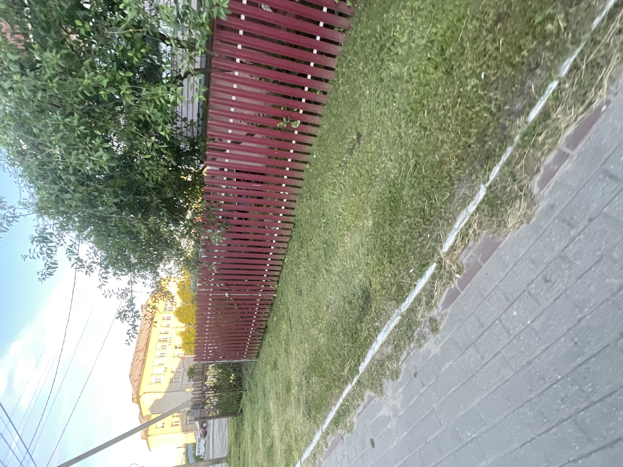 A sunlit, eye-level shot captures a quiet street scene in the city of Krasnye Baki, Russia. The foreground is dominated by a gray, paved sidewalk, bordered by a strip of mown grass and a vibrant red corrugated metal fence. Beyond the fence, a lush green lawn stretches towards a leafy tree. In the background, a large, light-colored building with a red roof stands prominently, its windows reflecting the bright sky. A few cars and a utility pole add to the urban landscape. The overall impression is one of peaceful, everyday life on a sunny day.