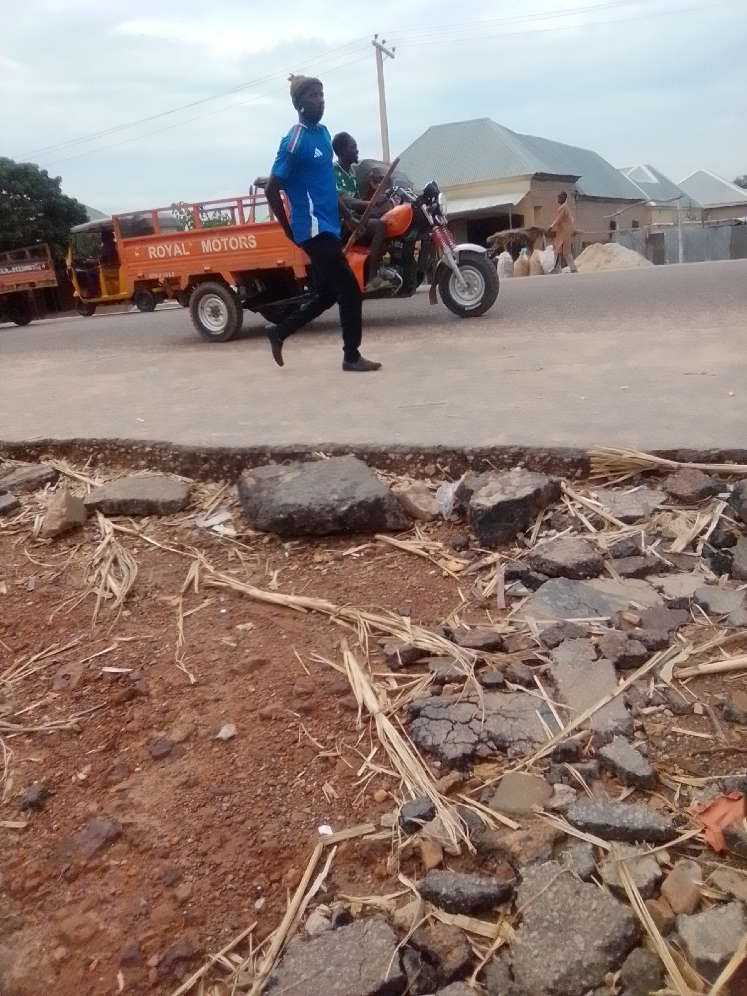 In Gombe, Nigeria, a person is walking on a street next to a three-wheeled truck with "ROYAL MOTORS" written on its side. Two other people are riding on a motorcycle behind the truck. The street is lined with buildings, some of which have corrugated metal roofs. The foreground of the image shows a rough, broken-up road surface with debris and dry grass. The sky is overcast.