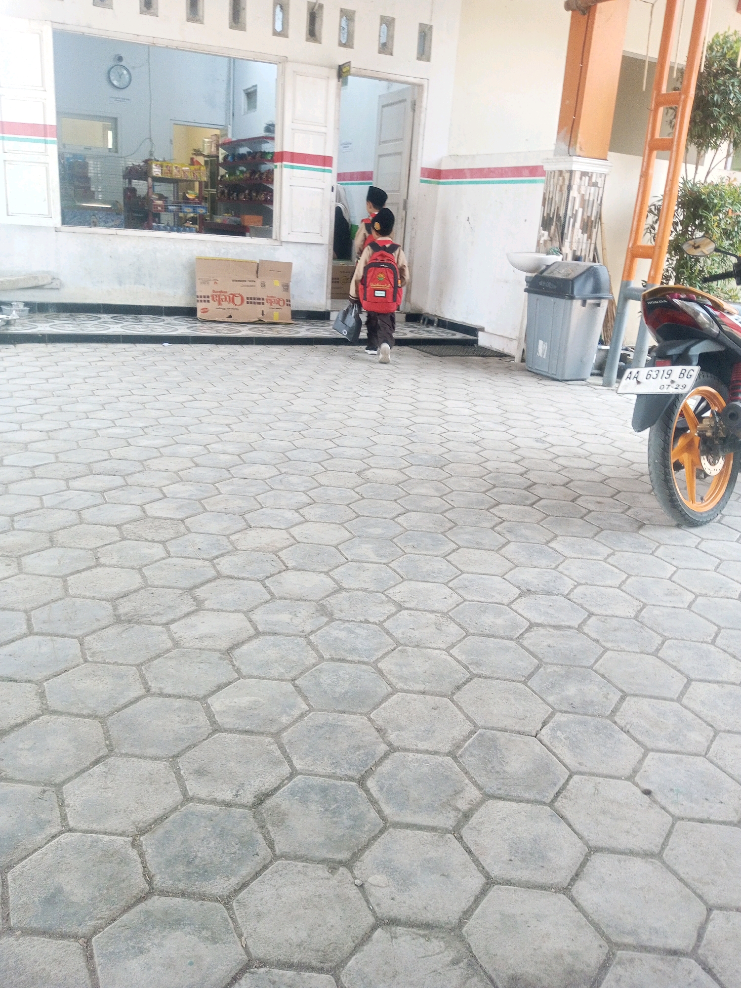 In Kalibeber, Indonesia, an outdoor scene captures a moment near a building with a large window revealing an interior with shelves stocked with items. Two children, one wearing a red backpack and the other's attire less distinct, are walking away from the building. A grey trash bin and a motorcycle with license plate "AA 6319 BG" are also present. The ground is paved with hexagonal concrete blocks.