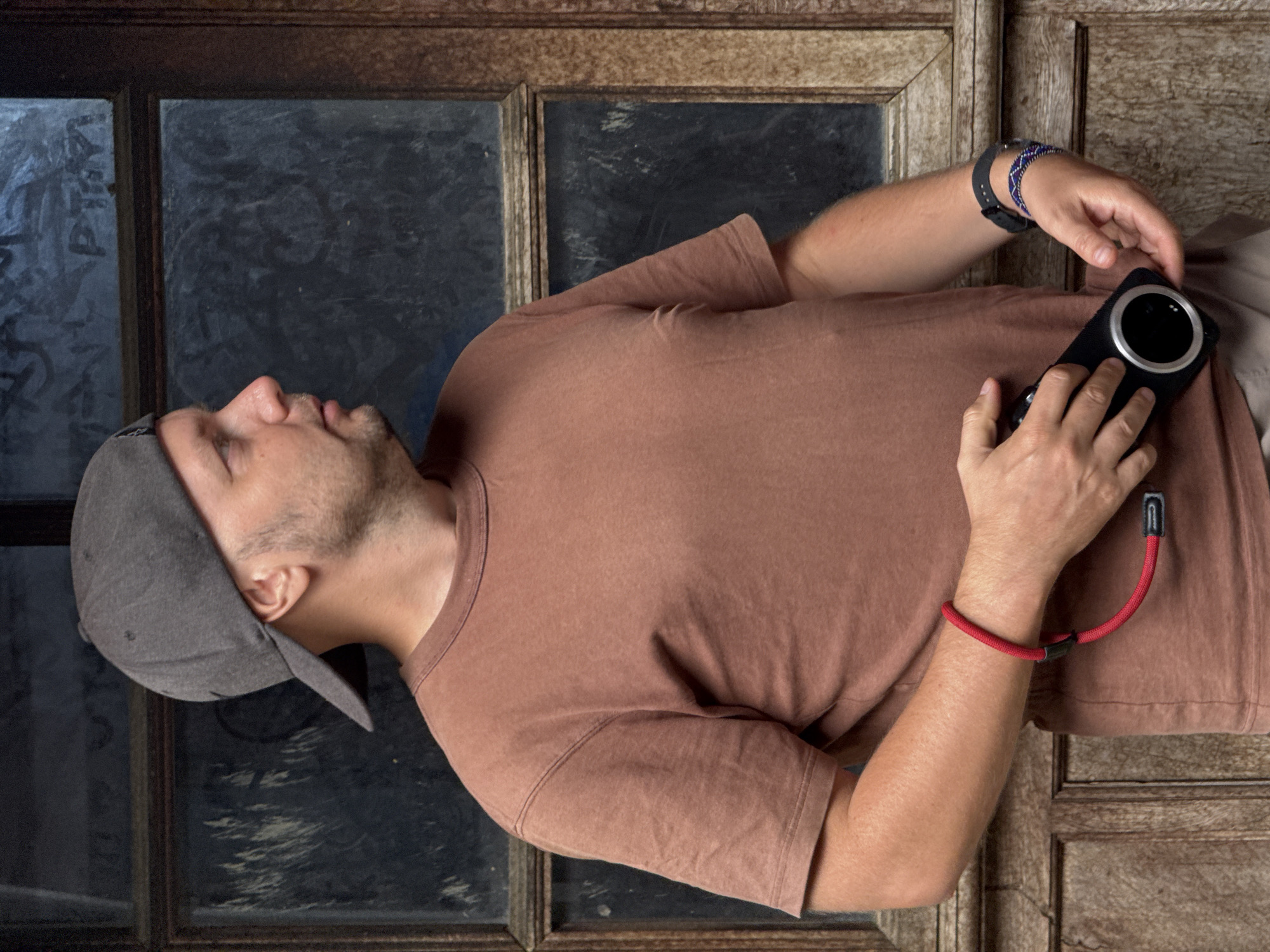 A man in a brown t-shirt and grey baseball cap is positioned in front of what appears to be an old wooden window frame. He holds a black smartphone with a large circular camera lens. The man has a dark watch and beaded bracelet on his left wrist and a red bracelet on his right. The man's face is tilted slightly up. The window behind him contains dark glass, possibly reflecting or obscured. The image is rotated; it would appear the man is lying down. The location of Sà Phìn Commune, Vietnam is not explicitly identifiable in the image.