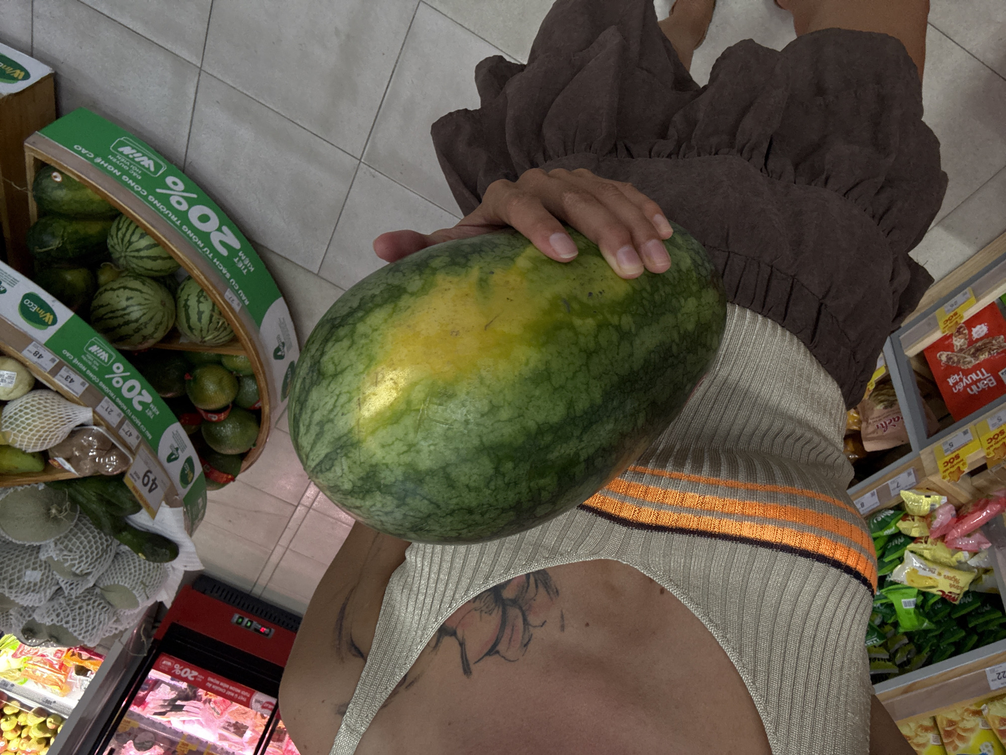 A person is holding a large watermelon in their right hand, in a grocery store. The person is wearing a beige knit top with orange stripes and a brown ruffled skirt. The watermelon is green with yellow markings. In the background, shelves are stocked with various food items and produce, including small watermelons in a wooden crate with a green price tag. The floor is tiled. The person's left arm is visible, with a tattoo on their upper arm. The image appears to be taken in a well-lit indoor environment.