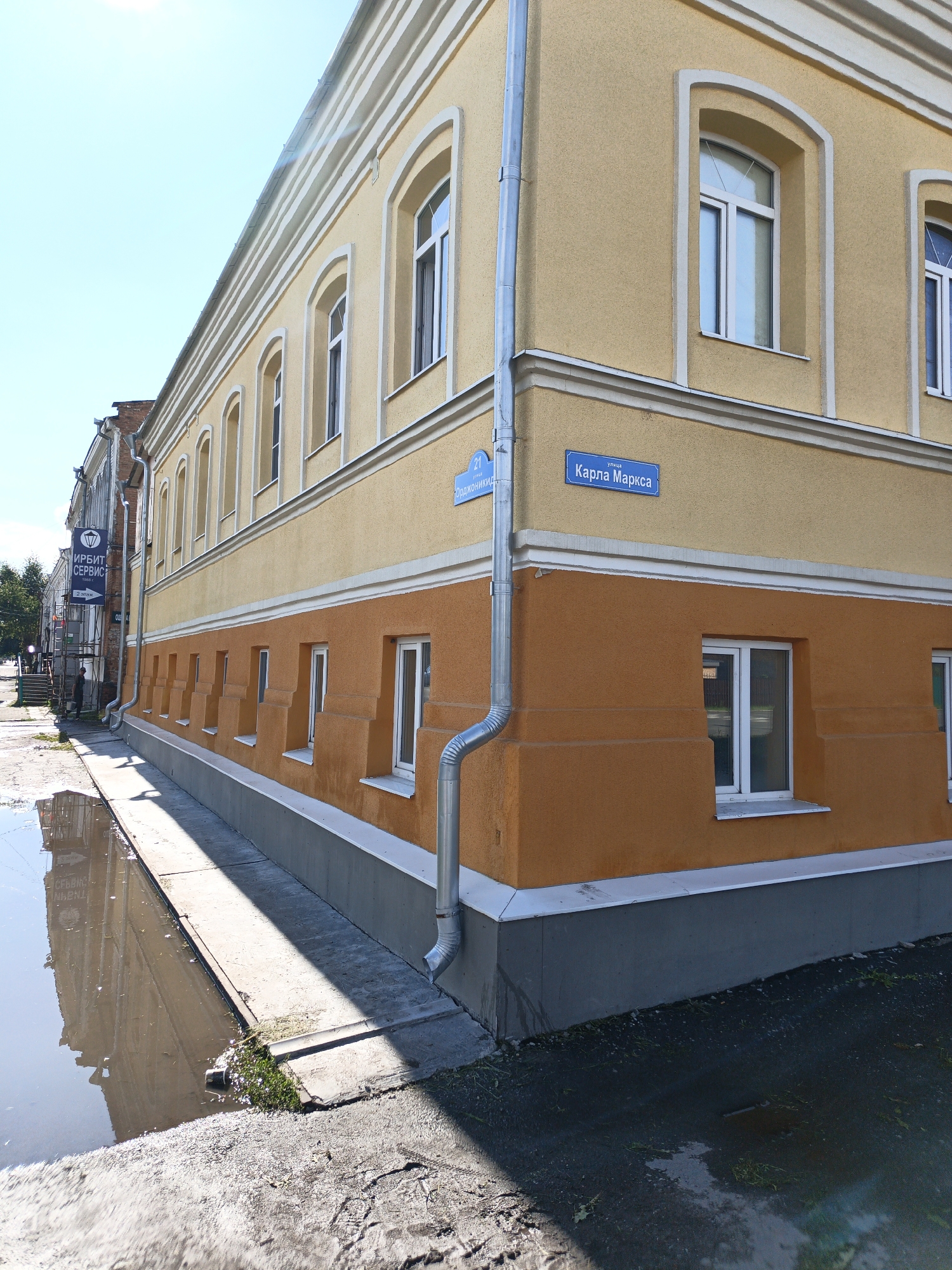 This image depicts a two-story building with yellow upper walls and orange lower walls, located on a street corner in Irbit, Russia. A blue street sign with white lettering indicates "улица Карла Маркса" (Karl Marx Street) on the yellow upper wall of the building. Another blue street sign, partially visible, reads "21 улица Орджоникидзе" (21 Ordzhonikidze Street). The building features arched windows on the upper floor and rectangular windows on the lower floor, all with white frames. A silver downspout runs vertically along the corner of the building. The street in front of the building is wet and reflects the building and sky, indicating recent rain. Some patchy grass is visible along the edge of the pavement. In the background, another building with a brick facade and a sign reading "ИРИБИТ СЕРВИС" is visible. The sky is clear and blue. No people are prominently featured in the image. The scene appears to be a typical urban streetscape on a sunny day after rain.