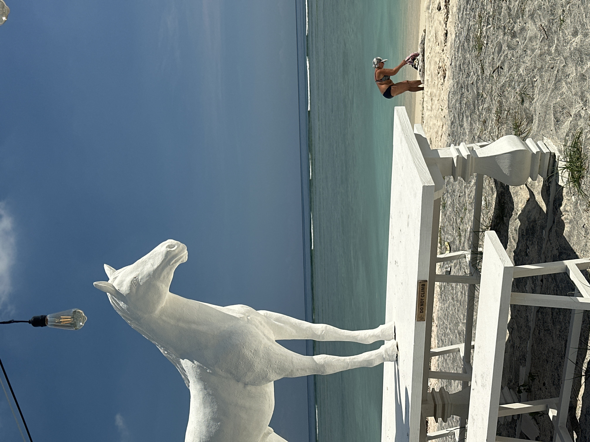 The image shows a white horse statue on a beach, in Unknown, Indonesia. The horse appears to be rearing up, facing the sky. A white decorative fence is nearby, with a small sign that reads "DO NOT CLIMB." In the background, a woman in a bikini and hat is standing on the sand near the water's edge, appearing to be interacting with an object on the beach. The water is turquoise, and the sky is blue with light clouds. A string of lights with a single bulb is visible to the left of the horse statue.
