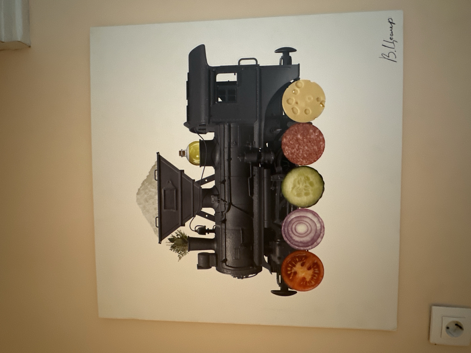 The image shows a painting of a black steam locomotive. The train's wheels are replaced by slices of food: a tomato slice, a slice of red onion, a slice of cucumber, a slice of salami, and a slice of cheese. The locomotive's tender is filled with salt, and a bottle of olive oil is on top of the boiler. A sprig of herbs is in the smokestack. The painting is signed in the lower right corner. The painting is hanging on a light brown wall, and a white electrical outlet is visible on the wall below and to the left of the painting. There is no specific information in the image to indicate its location in the city of Minsk, Belarus.