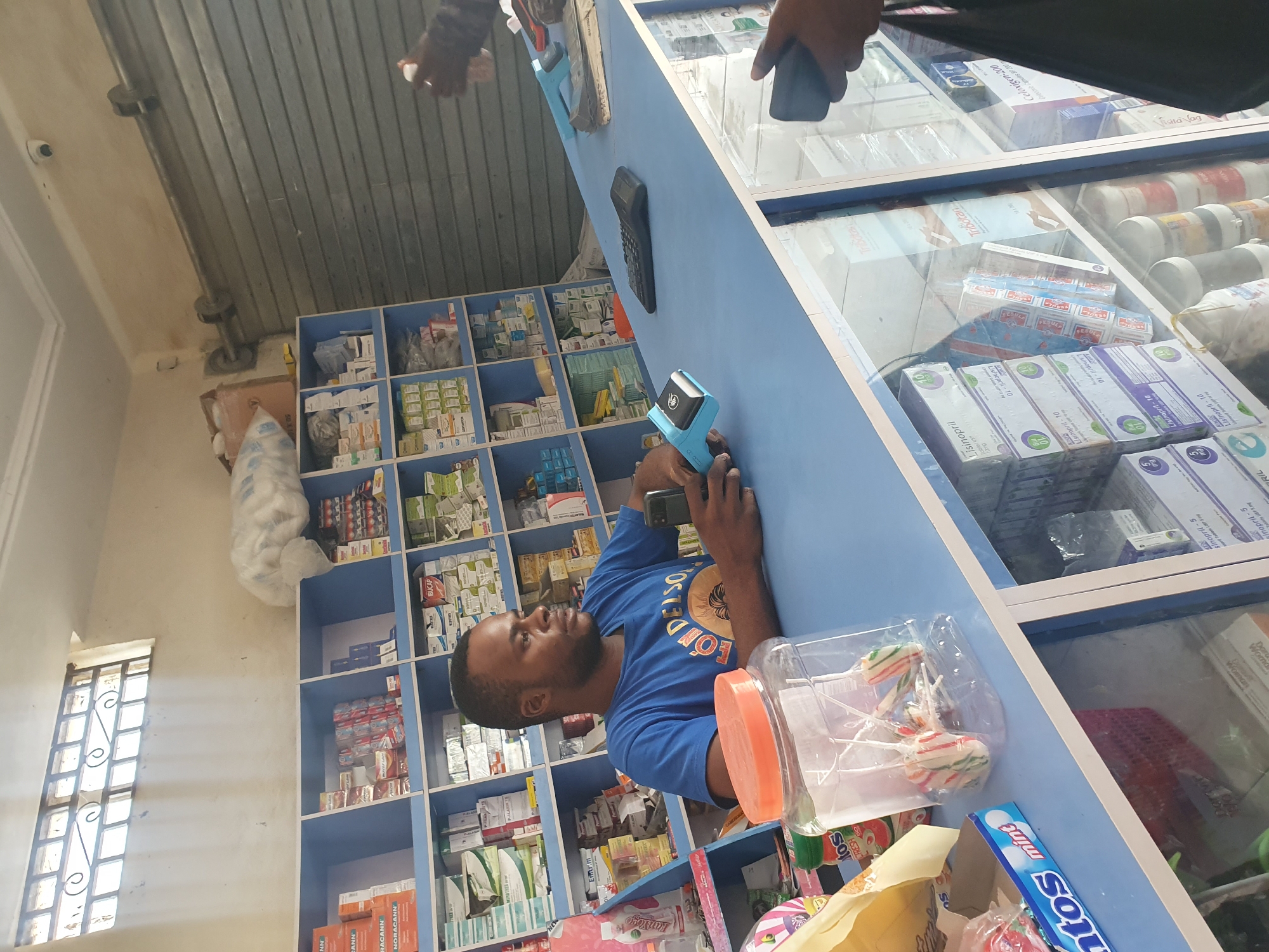 A man is standing in what appears to be a pharmacy in Gwagwalada, Nigeria, operating a mobile payment device to serve a customer. The pharmacy shelves behind him are filled with medications. In front of him, on the counter, there are lollipops, a Mentos box, and various pharmaceutical products behind a glass display case.