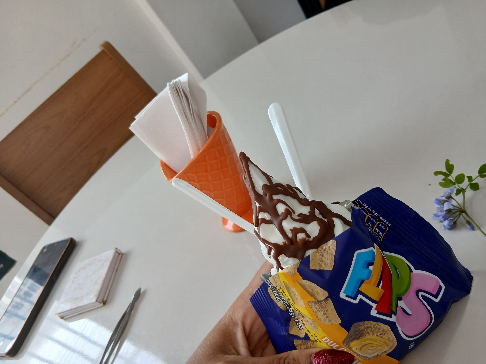 The image shows a hand holding an open bag of "Flips" brand corn snacks, with the bag partially obscuring a chocolate-covered ice cream cone. The cone is in an orange ceramic holder along with paper napkins and a white plastic spoon. Other objects visible on the white table are a cell phone, a small book, and a pair of tweezers. A small sprig of blue flowers is also present. The location, Juan Griego, Venezuela, is assumed based on the "Flips" branding which is common in South America.