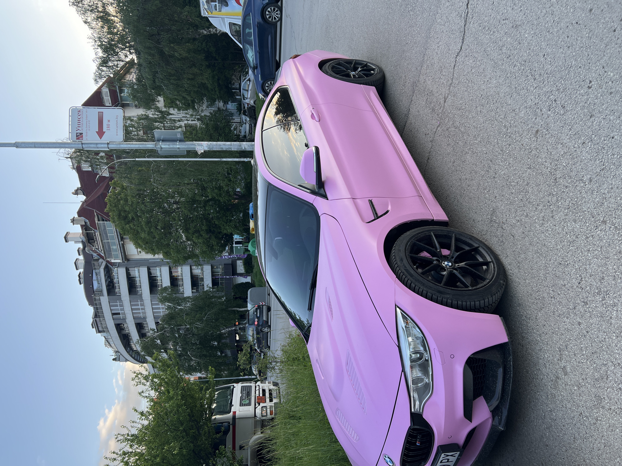 A pink BMW M4 coupe with black wheels is parked on the street in Sofia, Bulgaria. The car is parked on the right side of the road. In the background, there are several other cars, including a blue hatchback and a white delivery van. There is also a sign indicating directions to the "Унисекс" system, located 180 meters away. There are several buildings visible in the background, with a modern building on the left, and a building with a red roof further back. Trees line the street. The time of day appears to be late afternoon or early evening.