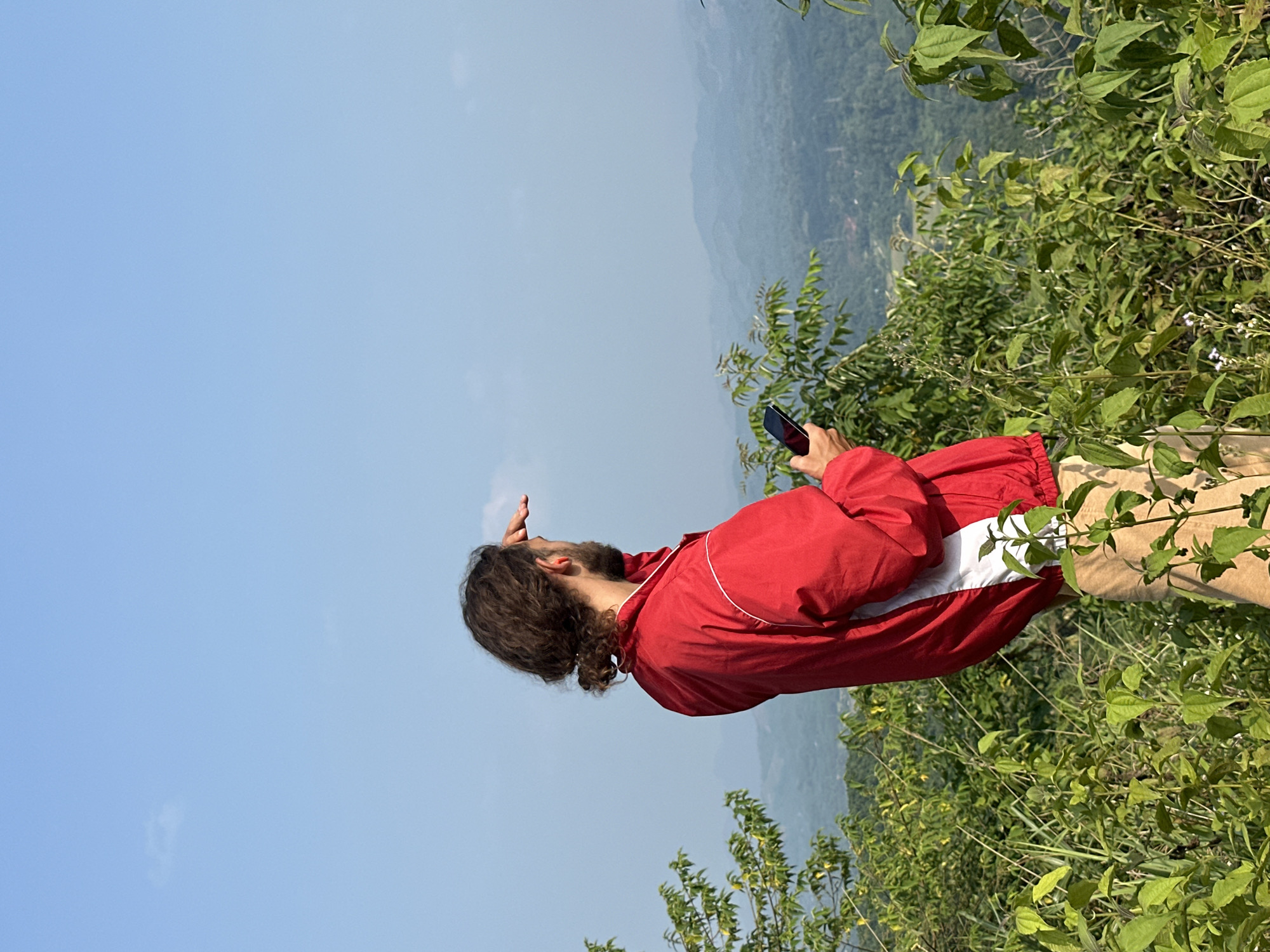 This image captures a person standing on a lush, green hillside, gazing upwards at the sky. The individual, seen from the chest up and slightly from behind, has long, dark, curly hair and a beard. They are dressed in a red jacket featuring a white horizontal stripe across the chest, and light-colored pants. Their right hand is raised, seemingly shielding their eyes from the sun, while their left hand holds a black smartphone, pointing it upwards as if to capture the view.
The scene depicts a clear, bright day over a mountainous landscape, consistent with the location of Tân Trịnh Commune, Vietnam. The foreground and midground are dominated by dense, vibrant green foliage and bushes, suggesting rich vegetation. In the background, distant mountains or hills recede under a wide expanse of clear, light blue sky. The image conveys a sense of outdoor exploration and appreciation for the natural environment. No visible text is present in the media.