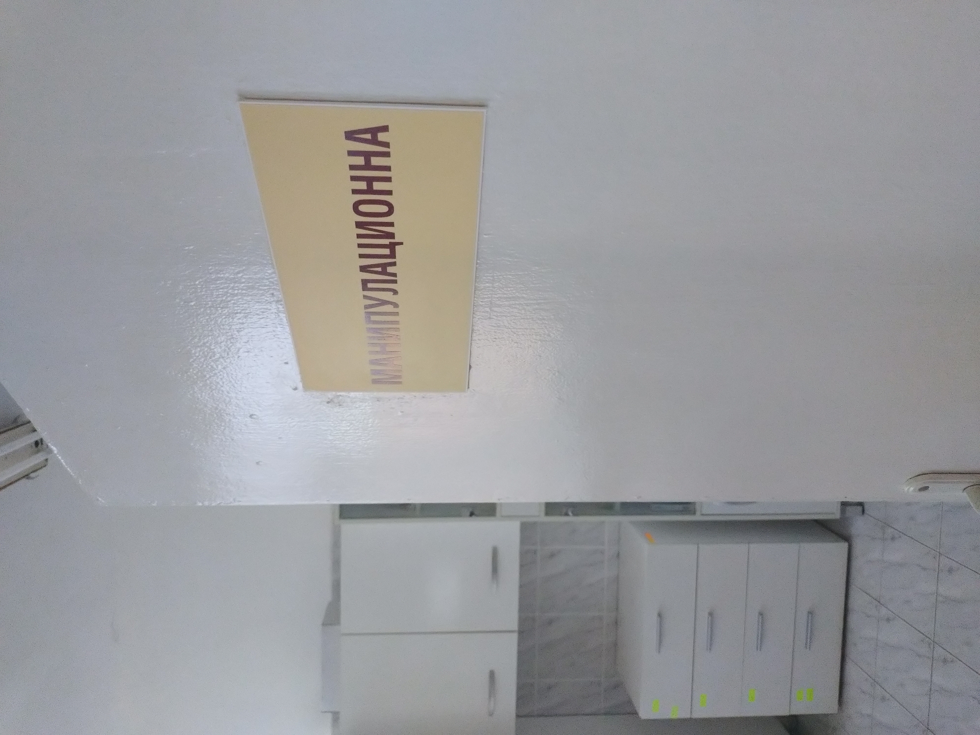 The scene appears to be the interior of a medical or healthcare facility. The environment is clean and sterile, suggested by the white walls, cabinets, and tile flooring. There is a sign in the upper portion of the image, with the text "МАНИПУЛАЦИОННА" written vertically on it. The word is in Cyrillic script. The location is likely Stara Zagora, Bulgaria. The overall environment indicates a medical setting.