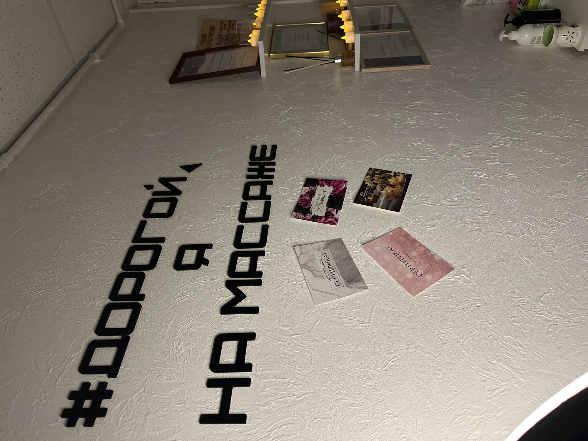 Here is a forensic description of the image. The location appears to be an interior space in the city of Unknown, Unknown, based on the text written in Russian.
The main subjects in the image are text and objects mounted on a white wall. The text, arranged in a vertical stack, spells out "#ДОРОГОЙ, Я НА МАССАЖЕ," which translates to "#DARLING, I AM AT A MASSAGE" in English. This text appears to be made of individual, thick, black letters and a hashtag symbol.
Also on the wall are four small cards or certificates. Each one has a unique design. Two of them are inscribed with the word "СЕРТИФИКАТ," which means "CERTIFICATE" in Russian.
On the upper part of the image are framed pictures and several bottles. To the extreme right, there are several bottles of what appear to be cosmetic products, including a white bottle with a pump dispenser.
The wall has a textured surface. Some of the objects appear to be attached to the wall, while others might be resting on a ledge. The lighting in the image suggests an overhead source, possibly a built-in lighting fixture. Part of a circular lamp is visible in the lower right corner.