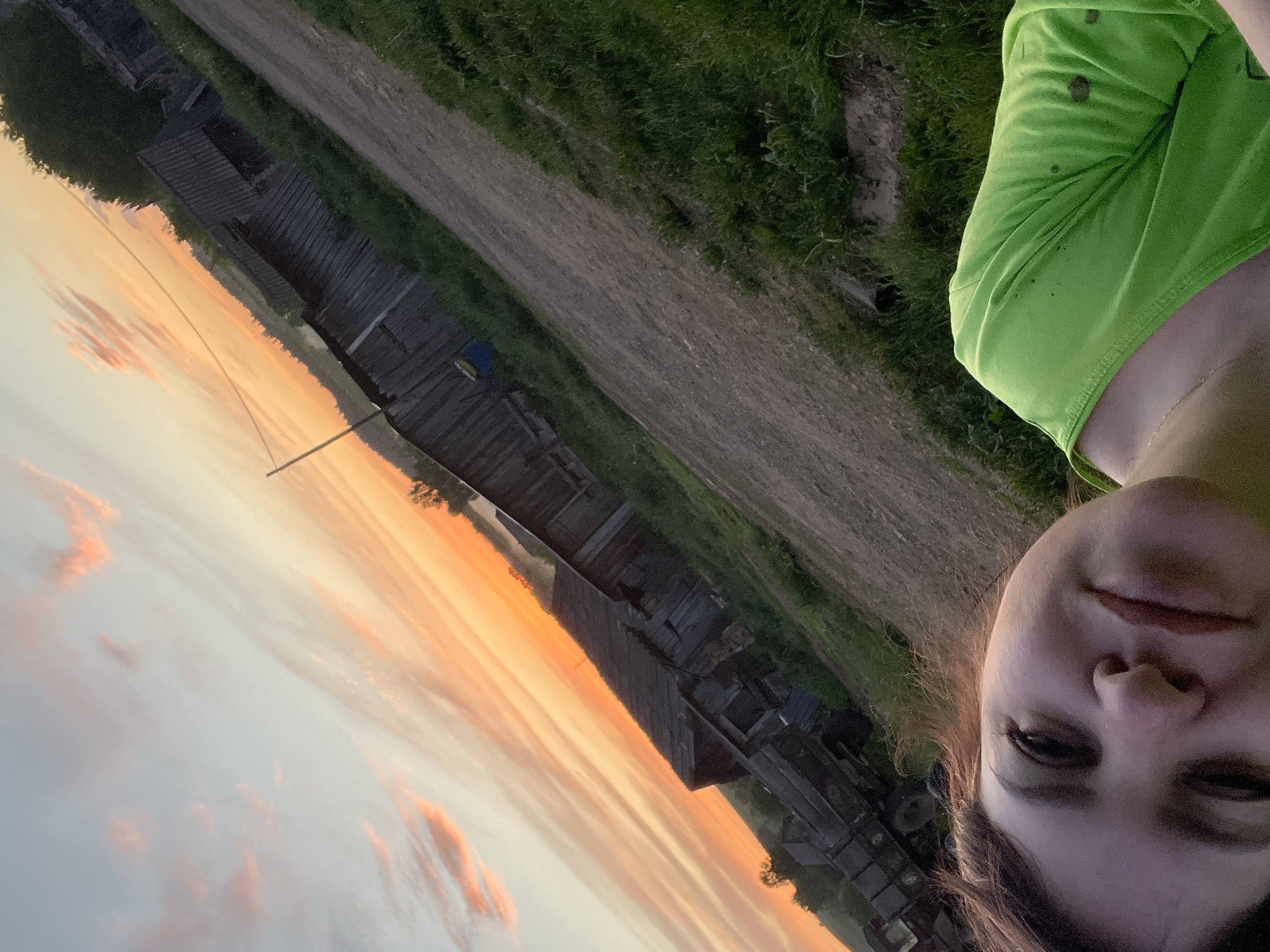 A woman is standing near a dirt road in Panteleevka, Russia, taking a selfie. To the woman's left is a row of aged wooden houses, while on the other side is a grassy area. The sky shows the colors of a sunset. The woman has brown hair and is wearing a bright green t-shirt.