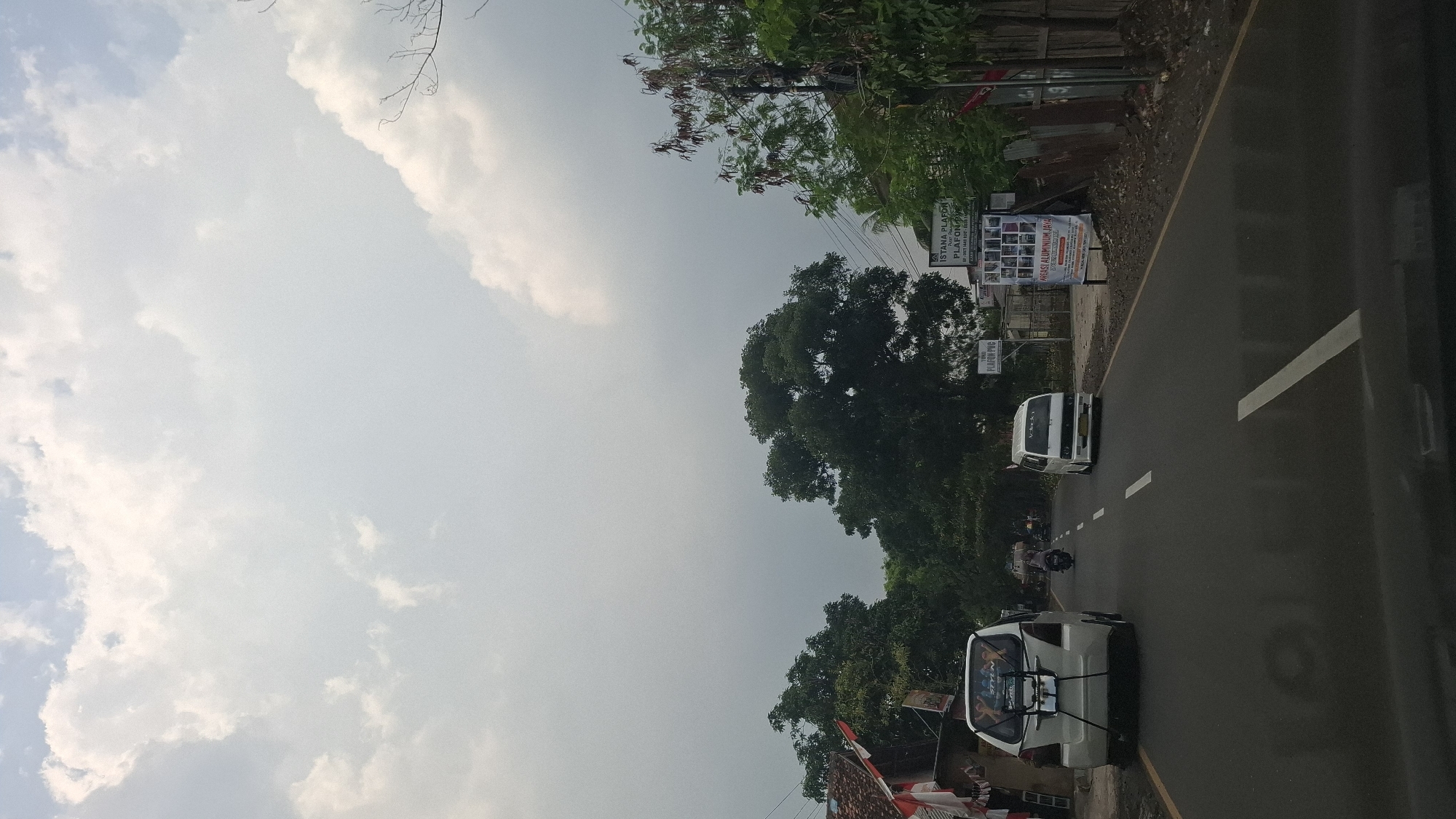 The image shows a road scene in Susukan, Indonesia, under a cloudy sky. A white van is parked on the left side of the road, with Indonesian flags attached to a nearby building. Further down the road, another white van is traveling, along with a motorcycle. On the right side of the road, there are several signs, including one that reads "ISTANA PLAFON" and another advertising aluminum services. Trees line both sides of the road, and the ground appears to be covered in gravel and dirt. The scene suggests a typical day in a rural or suburban area of Indonesia.