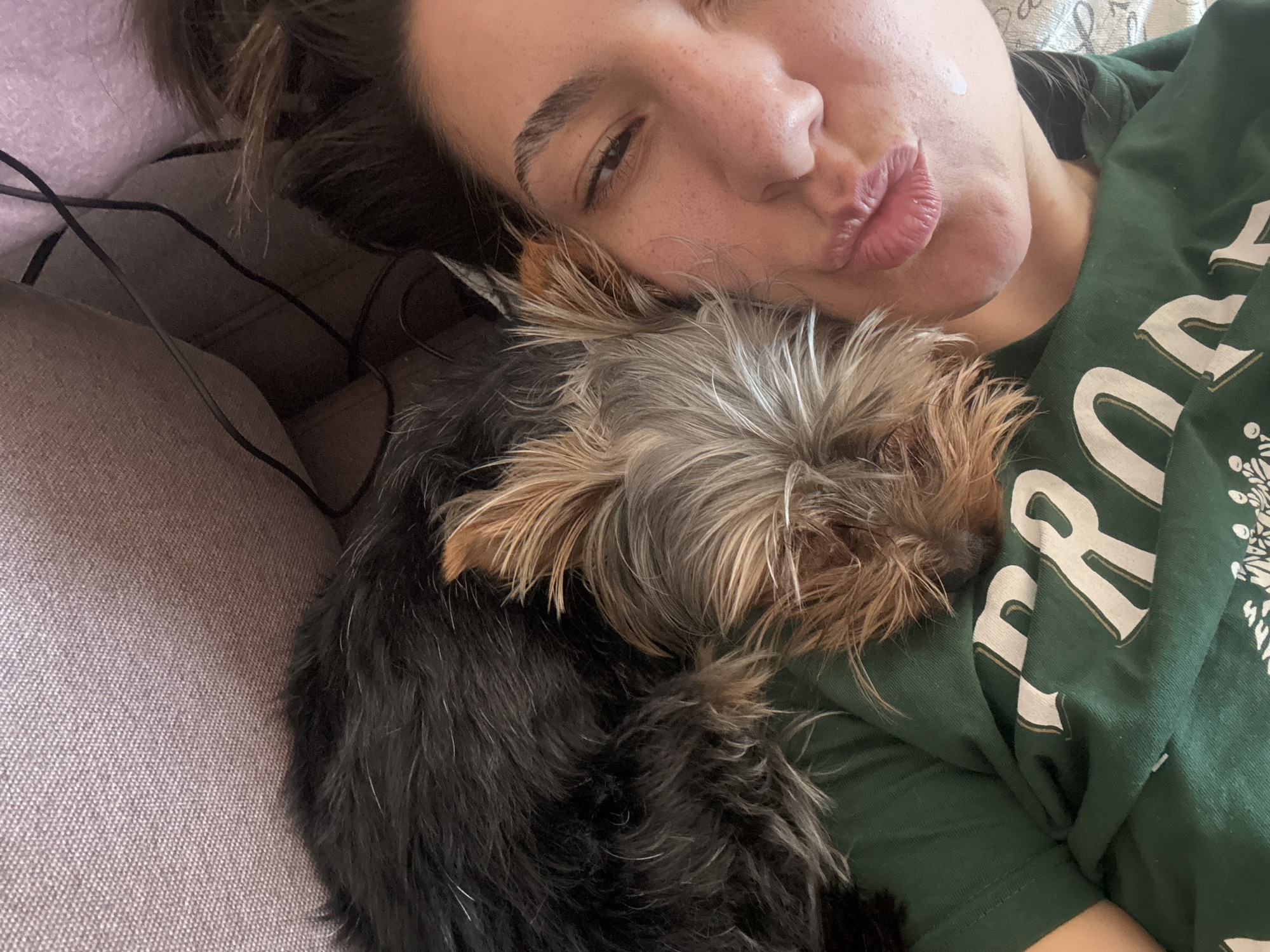 A woman is lying on a gray couch with a Yorkie dog nestled on her chest. She is wearing a green shirt with white lettering and appears to be making a kissing face at the camera. A white substance is visible on her cheek. The image does not contain information to determine a specific location in Varna, Bulgaria.