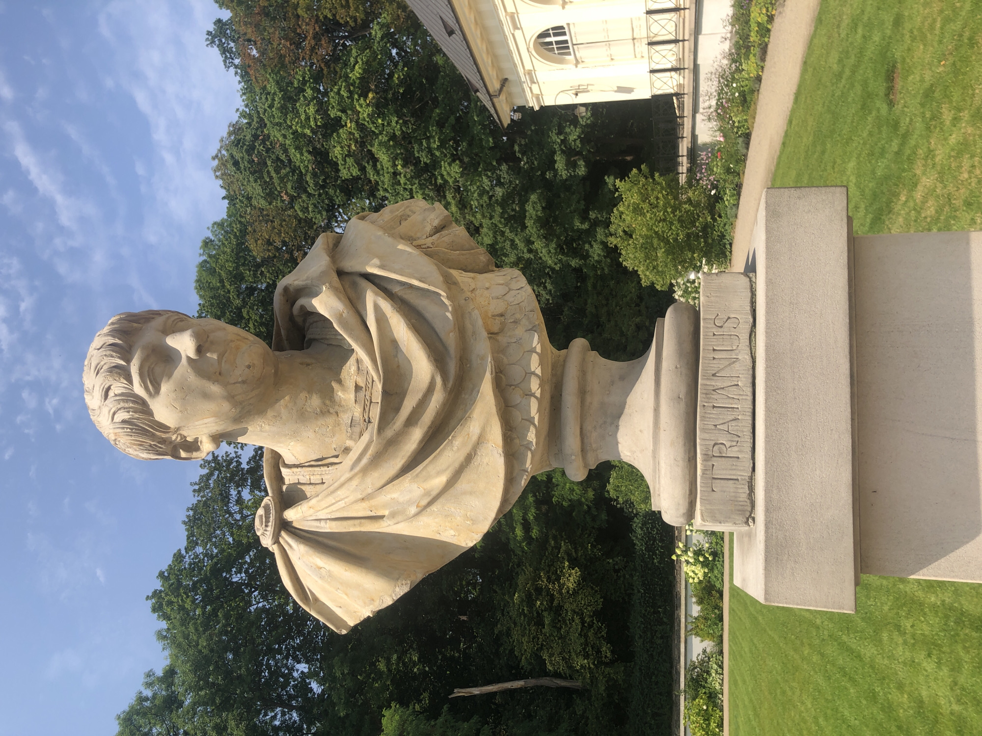 The image shows a stone bust of a Roman emperor, identified by the inscription "TRAIANUS" on its pedestal. The bust is made of light-colored stone and depicts a man with short hair, a toga draped over his shoulders, and a stern expression. The bust is placed on a multi-tiered pedestal, which is situated on a stone base.
The background of the image features a well-maintained garden with a manicured green lawn, flowerbeds with colorful flowers, and lush green trees. A path winds through the garden, and a classical-style building with arched windows is visible on the right side of the frame. The sky above is blue with scattered white clouds.
This bust is located in the Royal Baths Park (Łazienki Park) in Warsaw, Poland. The park is a historical landmark known for its beautiful gardens, palaces, and sculptures. The bust of Trajan is part of the park's collection of Roman emperors' sculptures, arranged around the amphitheater.