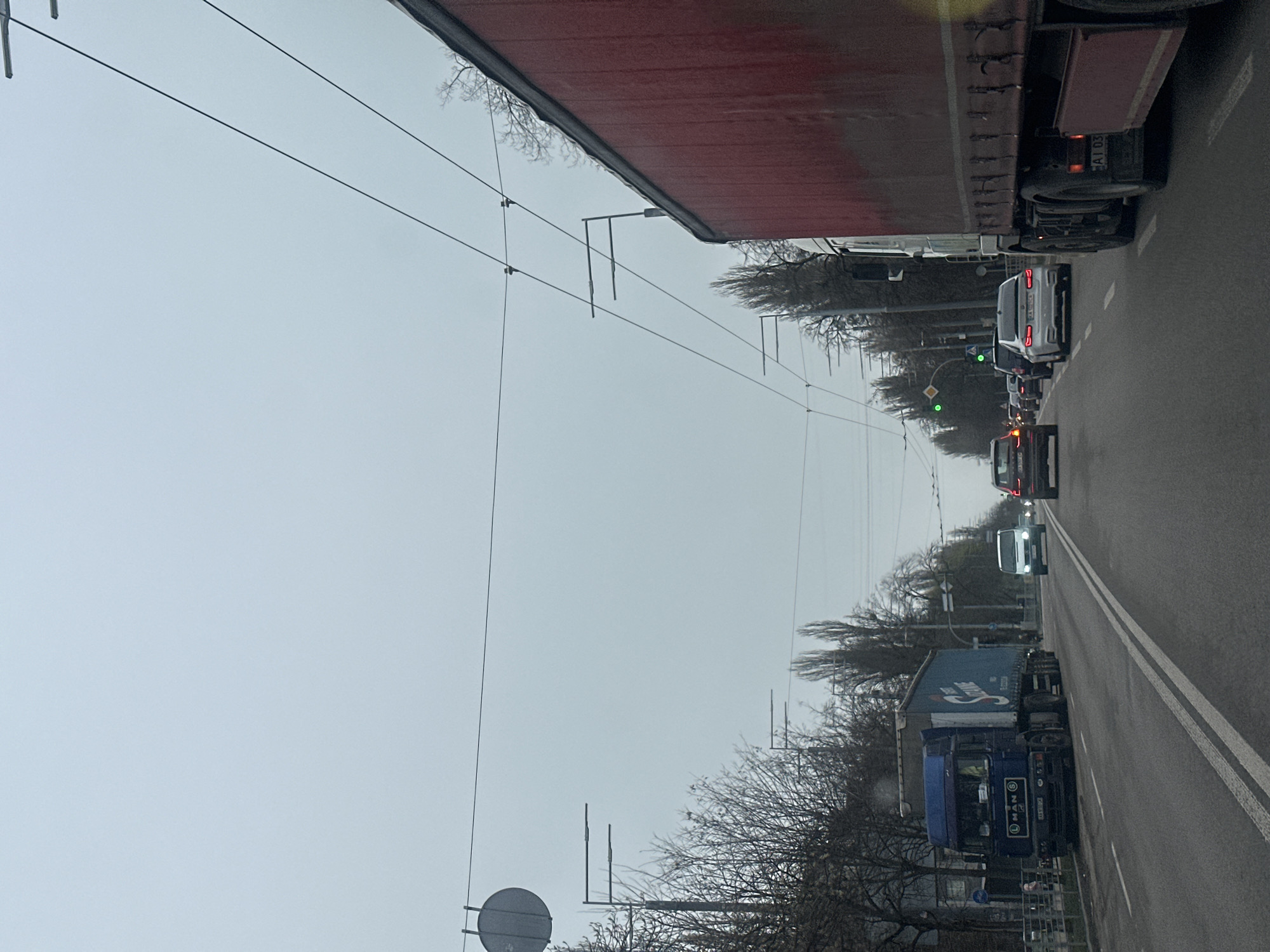 The image shows a street in Kyiv, Ukraine, with heavy traffic. The sky is overcast. A large red truck is on the right side of the road. Several cars and a blue MAN truck are in the opposite lane. Traffic lights are visible in the distance, with some showing green. Bare trees line the street. Overhead power lines cross the road. The image is taken from the side of the road.
