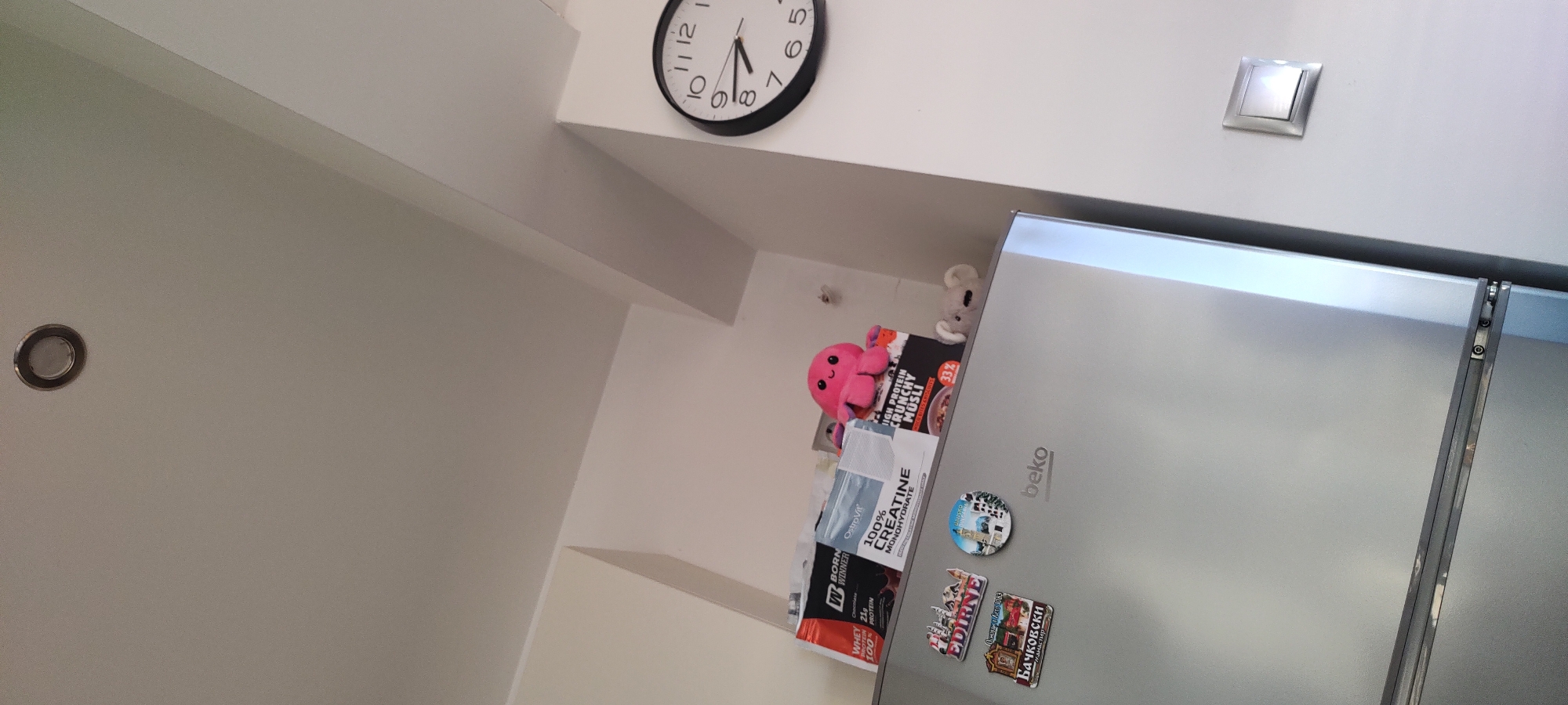 Here is a description of the image:
The image shows a portion of a room, likely a kitchen, with a focus on the upper area of a white wall. A black round clock hangs near the ceiling, indicating the time is close to 7:05. A light switch is mounted on the wall to the right.
Below these fixtures, a refrigerator is visible. Its surface is adorned with various items, including a pink stuffed octopus, a box of "HIGH PROTEIN CRUNCHY MÜSLI", a container of "OstroVit 100% CREATINE MONOHYDRATE", and a packet labeled "WWD BORN WINNER". Several magnets featuring images, including one that says "EDIRNE" and "Бачковски", are also attached to the refrigerator door. A "beko" logo is visible on the refrigerator.
