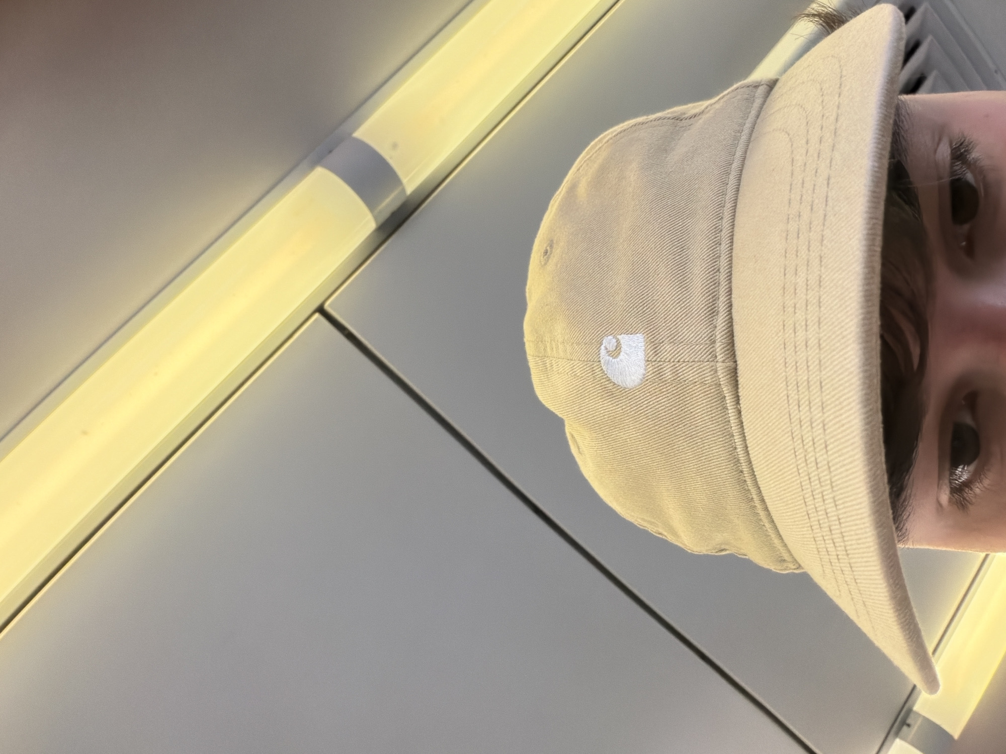 A person wearing a beige Carhartt baseball cap is looking directly into the camera. The person has dark hair and brown eyes. The background is a ceiling with fluorescent lighting. The setting appears to be an indoor space, possibly an airplane or train carriage. There is no indication of the location being in Lisbon, Portugal.