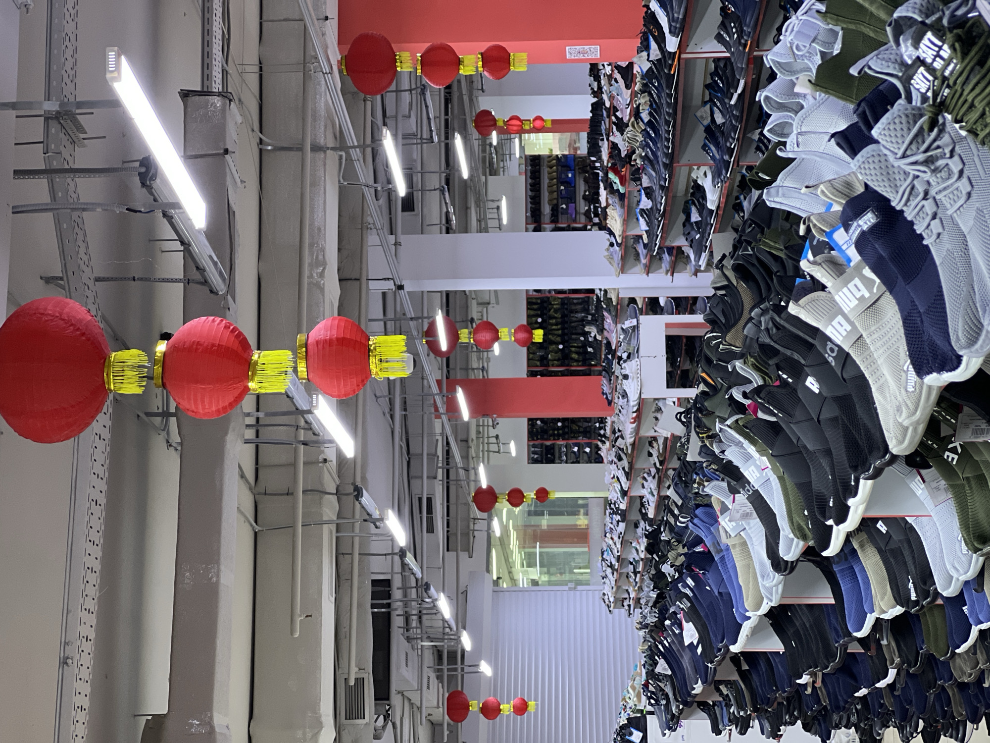The image depicts a retail environment, likely a shoe store, with numerous pairs of athletic shoes displayed on tiered shelving units. The store is brightly lit by fluorescent ceiling lights and appears to be indoors. Red Chinese lanterns are suspended from the ceiling at various heights, suggesting a festive or decorative theme. The shelves are filled with a variety of shoes, predominantly sneakers, in a range of colors and styles. Some shoes are displayed in boxes, while others are placed directly on the shelves. In the background, more shelving units and potentially other sections of the store are visible, along with what appears to be a rolling metal door or gate on the left side. The image lacks any discernible elements that would place it specifically in Krasnodar, Russia, or any other particular city. There are no people visible in the image.