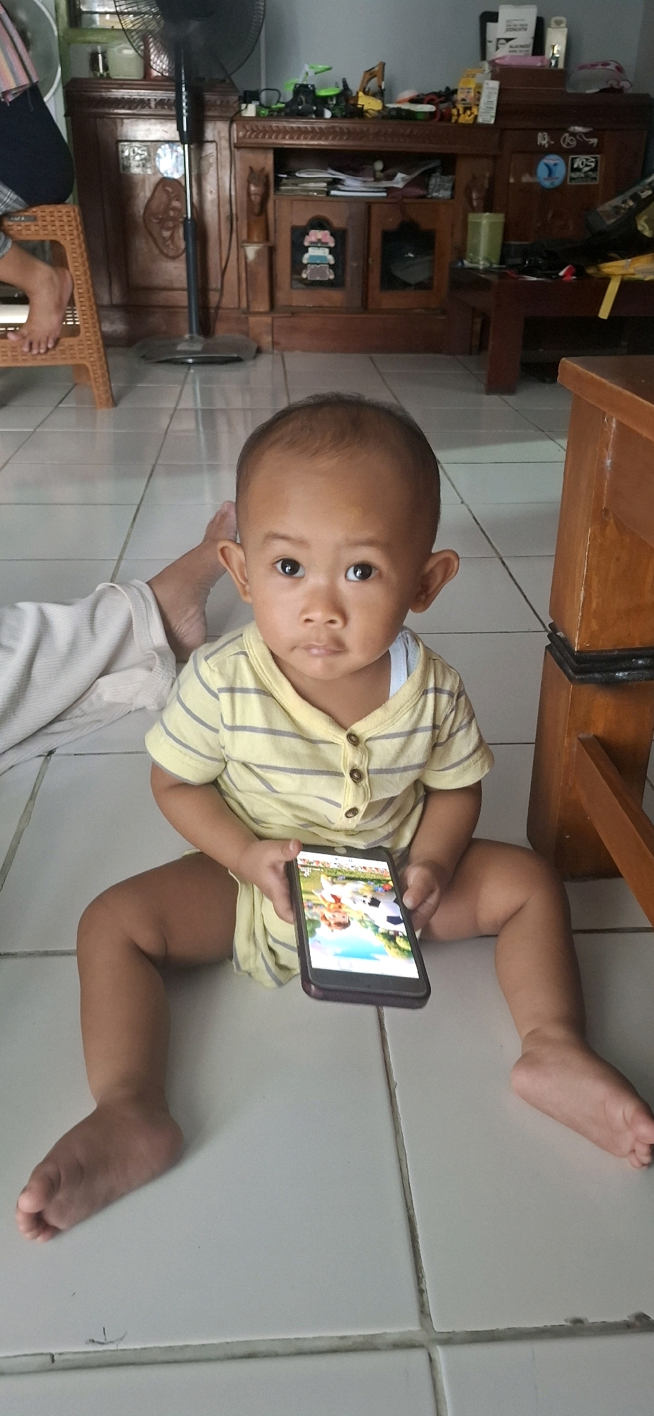 A young child, appearing to be a toddler, sits on a tiled floor in Desa Gagaksipat, Indonesia, holding a smartphone. The child is wearing a yellow and grey striped t-shirt and is looking directly at the camera with a neutral expression. The smartphone displays an image of animated characters playing soccer. To the left of the child, a person's leg and foot are visible, partially covered by white fabric. In the background, a wooden cabinet filled with various items, including toys and decorative objects, is visible. A black floor fan stands behind the cabinet. The overall scene suggests a domestic setting.