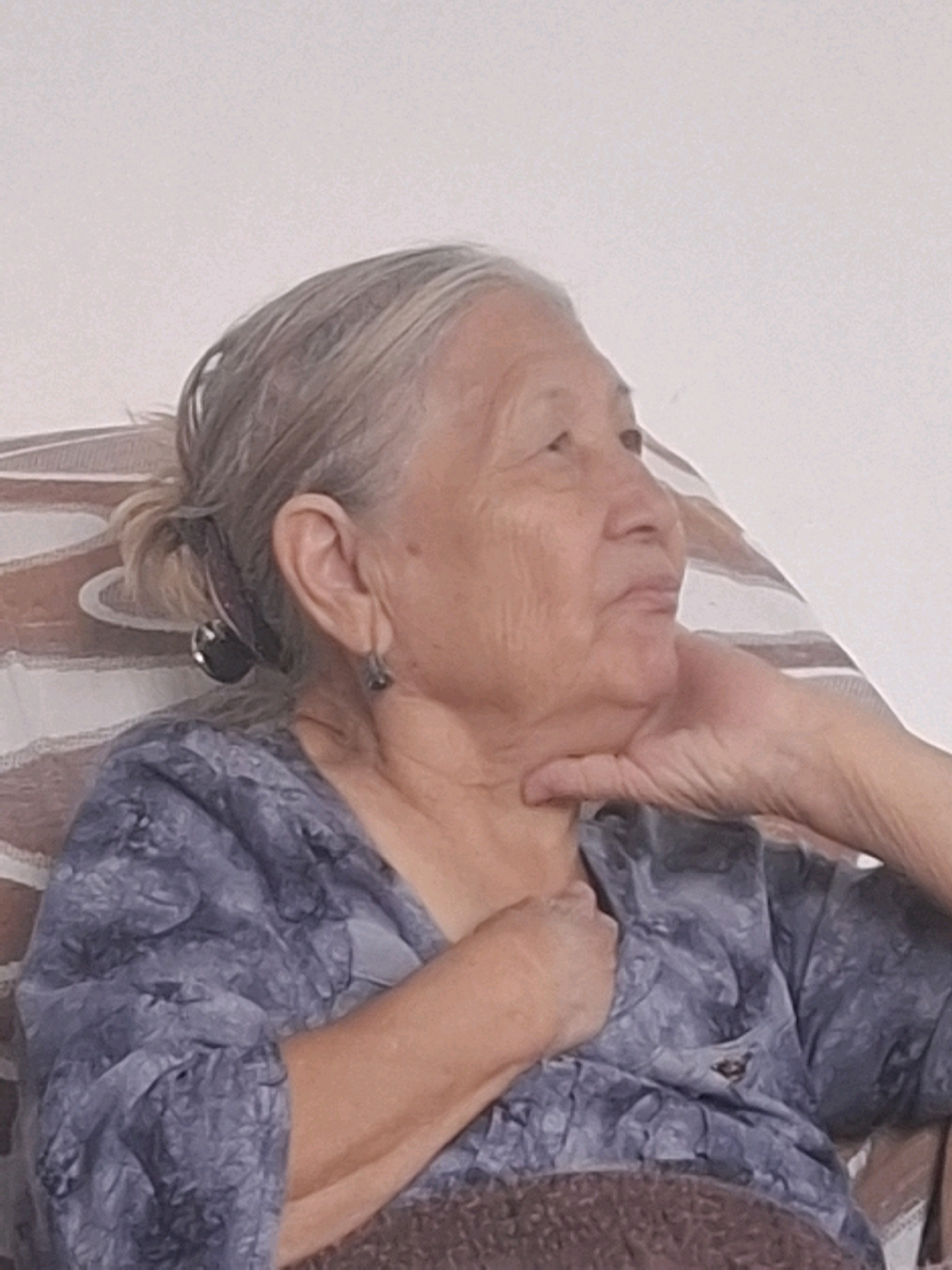 An elderly woman with graying hair tied back in a bun is captured in a medium shot. She is seated, leaning her head on her hand and gazing upwards and to the right. Her attire consists of a blue, patterned shirt. The background suggests an indoor setting, possibly a living room, with a patterned sofa and a plain wall. The lighting is soft and diffused, creating a gentle ambiance. The image lacks any contextual clues to pinpoint its specific location within Almaty, Kazakhstan.