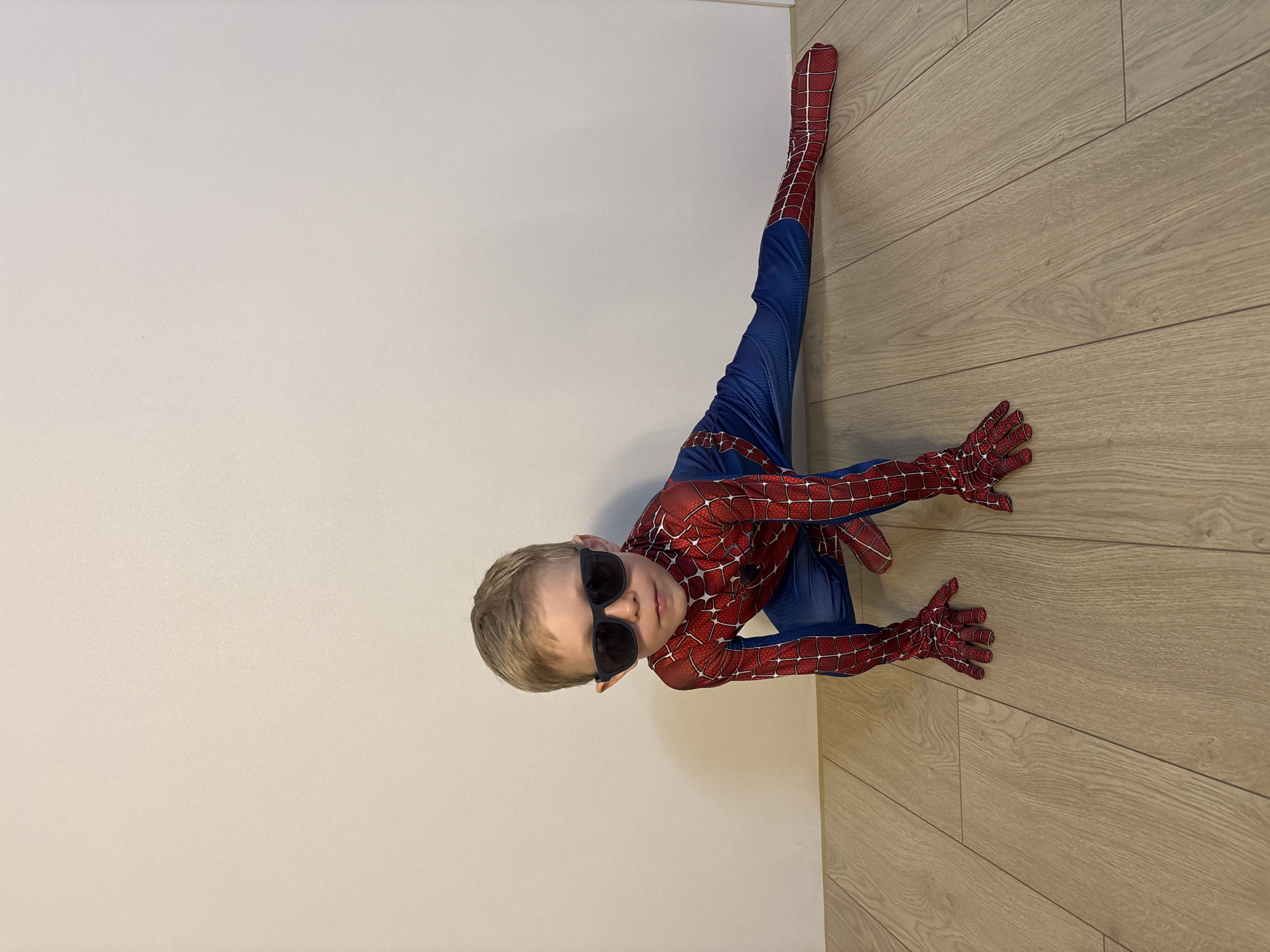 A boy in a Spider-Man costume is positioned in the corner of a room. The room has a light-colored wooden floor and a light beige wall. The boy is wearing black sunglasses. One leg is extended up the wall, while his hands are placed on the floor.
The location is not discernible from this image. It is not possible to determine the location, nor if it is in Chaikovsky, Russia.