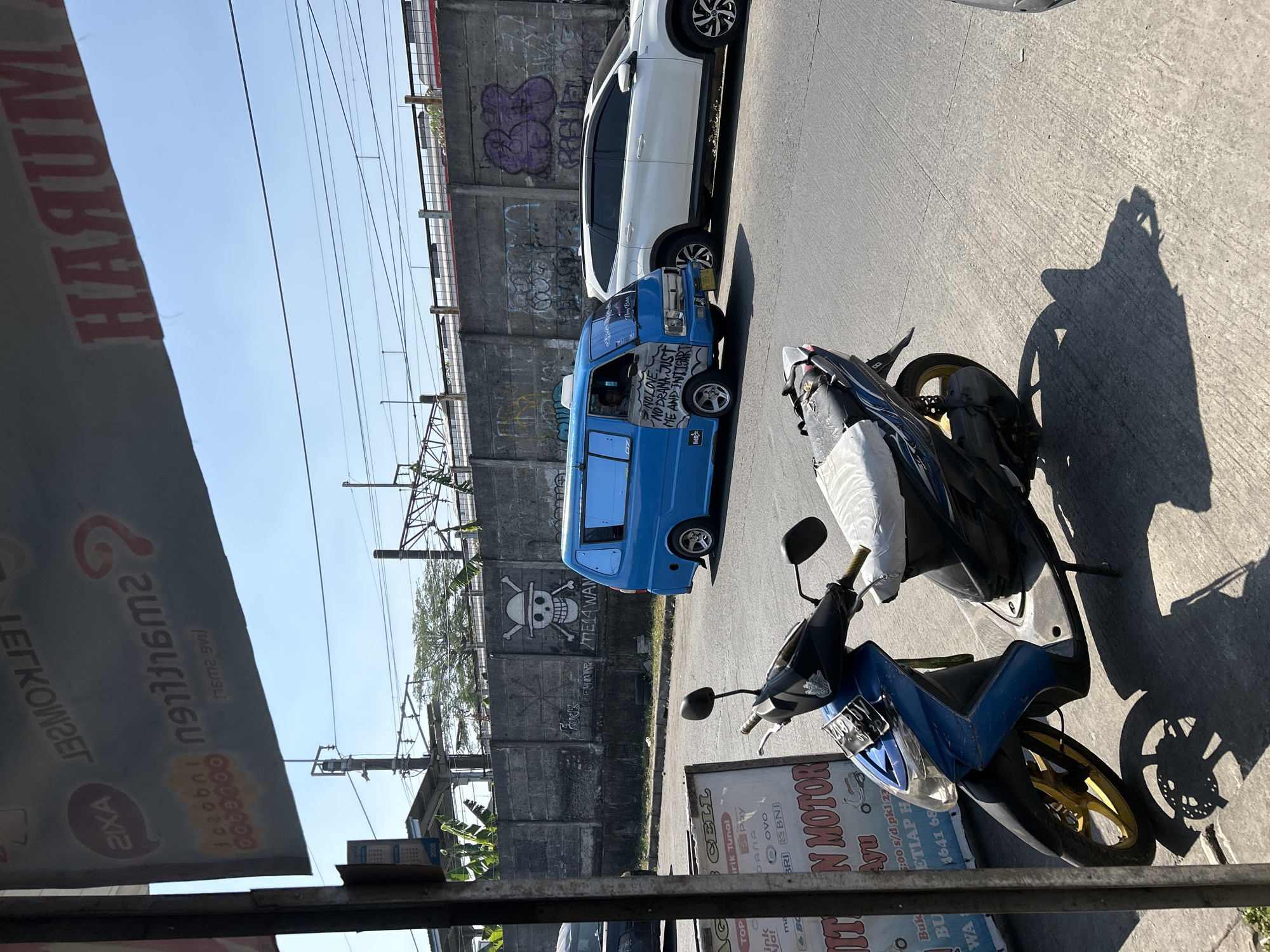 A blue van with the text "NO LOVE NO DRAMA JUST ME AND INTIGART" written on its side is parked on a street in Depok, Indonesia, next to a white SUV. In the foreground, a blue motorcycle is parked on the sidewalk, partially covered with a white sheet. Behind the vehicles, a concrete wall is covered in graffiti, including a skull and crossbones image. Overhead, power lines and train tracks are visible, with a train in the distance. The sky is clear and blue. A sign in the foreground advertises "MITRA MOTOR".