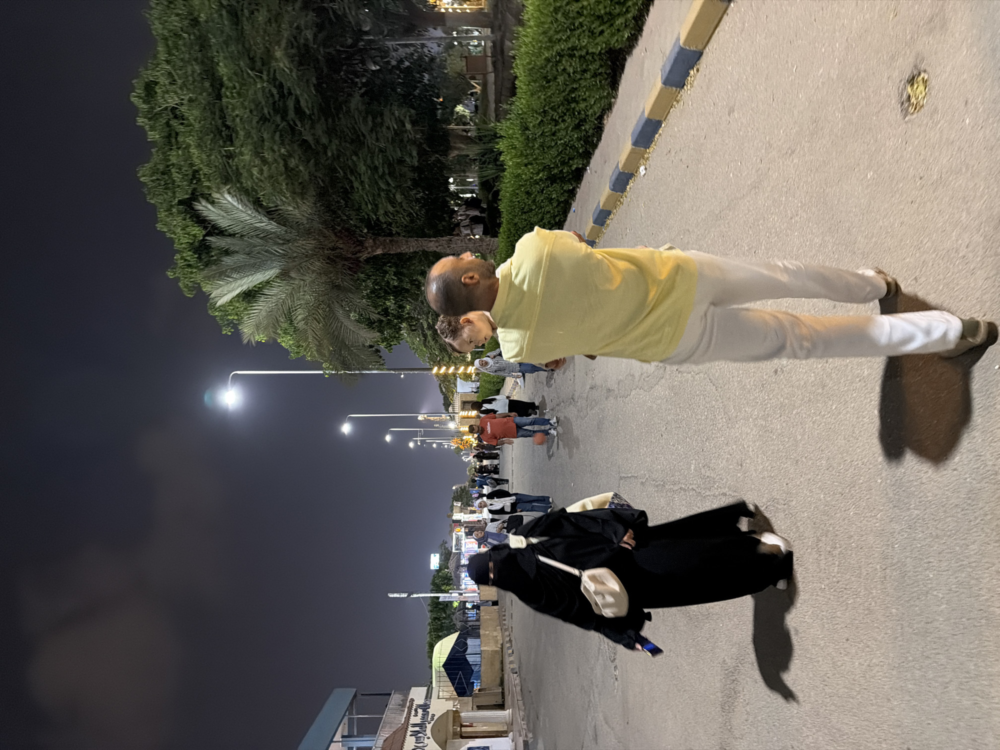 The image captures a nighttime scene on a paved street, likely in the city of 6th of October, Egypt, given the context. The street is lined with sparse palm trees and dense green bushes. In the foreground, a man with a bald head, wearing a yellow short-sleeved shirt and white trousers, carries a young child on his left hip, looking to the right. Behind him, a woman dressed in a black abaya and hijab, with a light-colored bag slung over her shoulder, walks away from the camera, holding a dark object in her right hand. Several other pedestrians, including a man in a red t-shirt and people in casual attire, are visible further down the street, illuminated by streetlights. Buildings with signs, some featuring Arabic script, are present on the left side of the street. The sky is dark with scattered clouds.