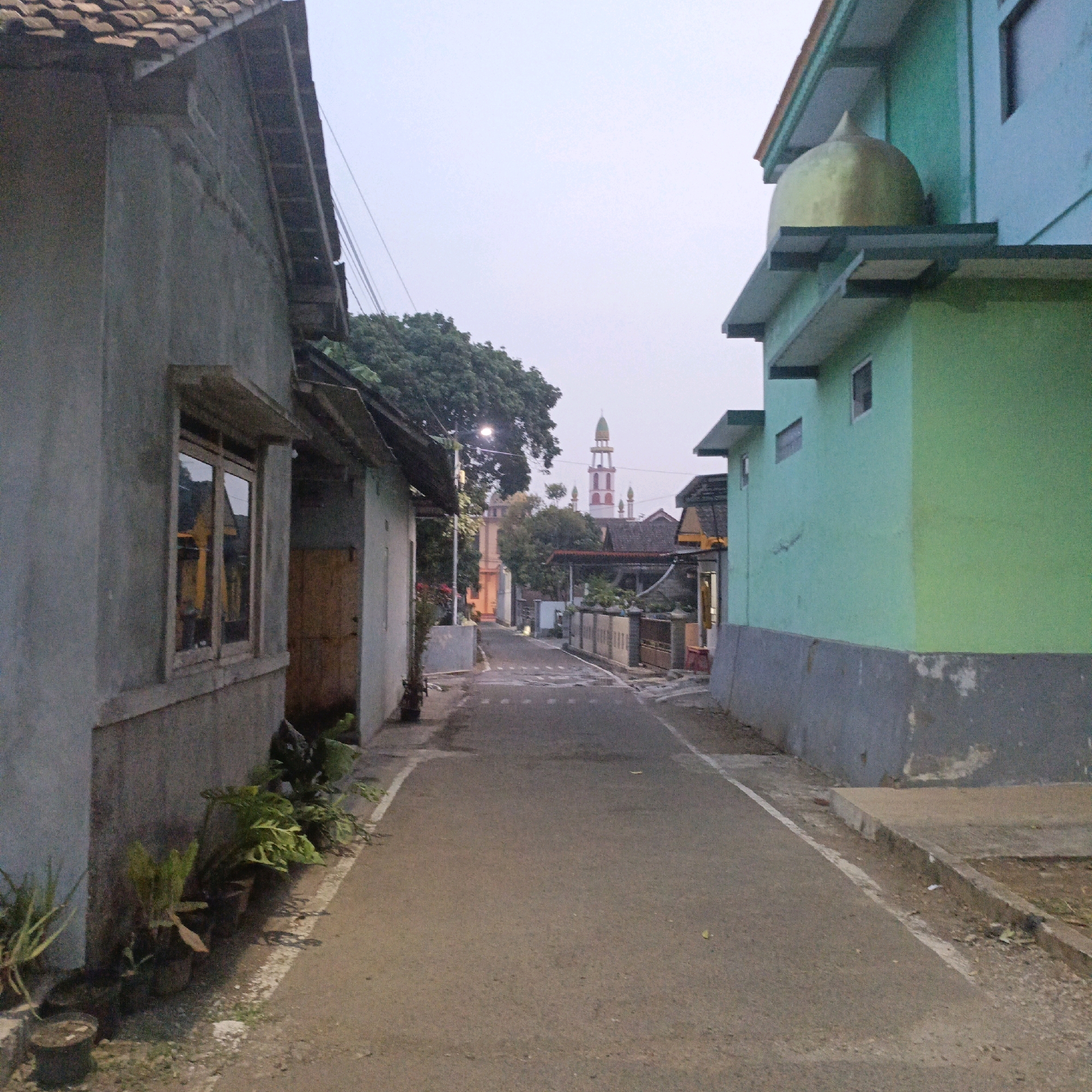 The image depicts a narrow asphalt street in a residential area, likely in Salatiga, Indonesia, with buildings lining both sides. The sky is overcast, suggesting dawn or dusk. On the left, a gray, two-story building with a tiled roof and a visible window frame is prominent. Plants in pots are arranged along the base of this building.
On the right, a light green two-story building with a gray concrete foundation is visible. A golden dome-shaped structure, possibly part of a smaller mosque or religious building, is situated on the upper level of this structure.
Further down the street, a tall mosque minaret with a red and white striped design and a green dome is visible against the pale sky. Other buildings with red tiled roofs and various architectural styles are scattered throughout the background. The street is sparsely populated, with no people visible in the frame. The overall scene conveys a quiet, everyday atmosphere of a local Indonesian neighborhood.