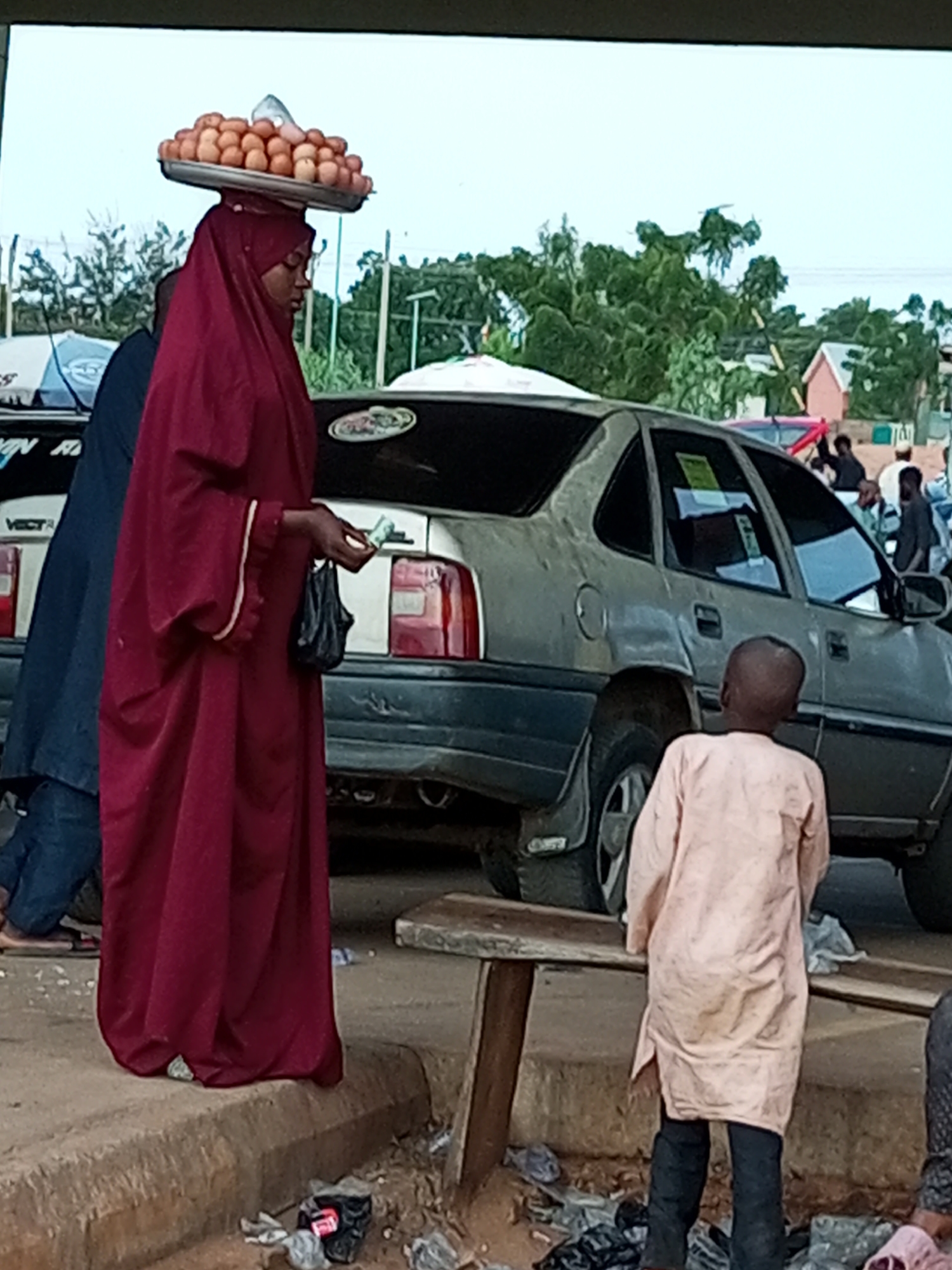 In the city of Gombe, Nigeria, a woman in a maroon hijab is seen balancing a tray of eggs on her head. She is standing next to a light-colored, older model sedan. To her right, a young boy in a pale pink tunic and dark pants faces away from the camera, looking towards the car. The ground is dirt and debris, with some trash visible near the curb. In the background, there are trees, buildings, and other people. The overall scene suggests a street market or a roadside scene.