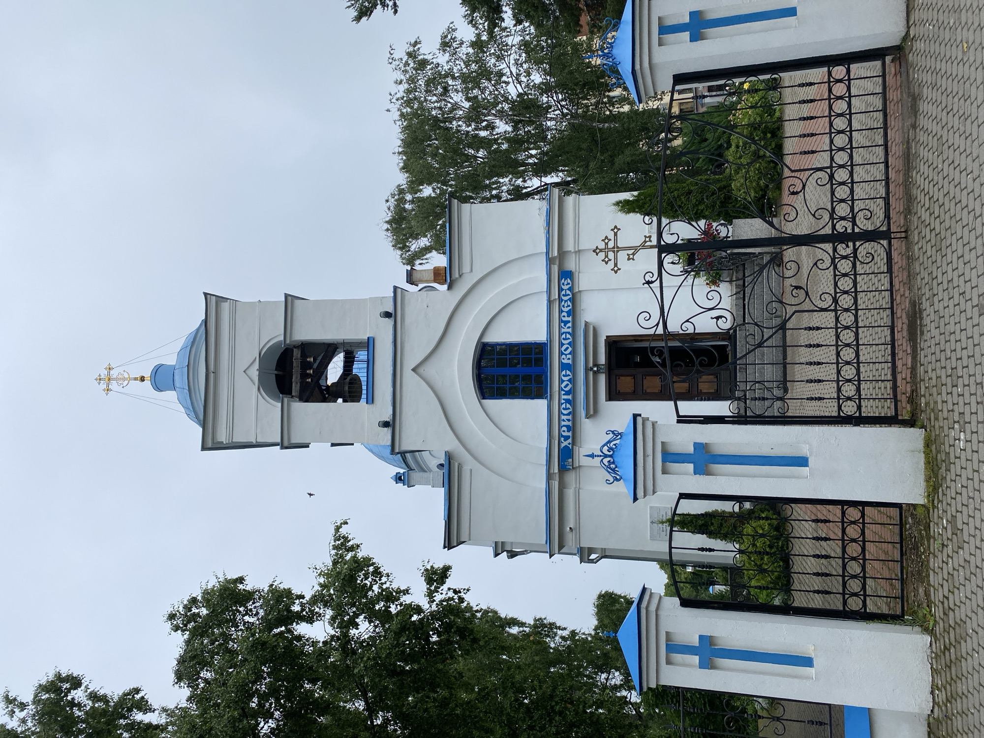 The image shows the exterior of an Eastern Orthodox church in Priozersk, Russia. The church is white with blue accents on its domes, windows, and decorative elements. A bell tower with a golden cross tops the building. The church is surrounded by a black wrought-iron fence with white pillars adorned with blue crosses. Trees line the property, and the sky is overcast. The phrase "ХРИСТОС ВОСКРЕСЕ" (Christ is Risen) is written in blue letters above the entrance.