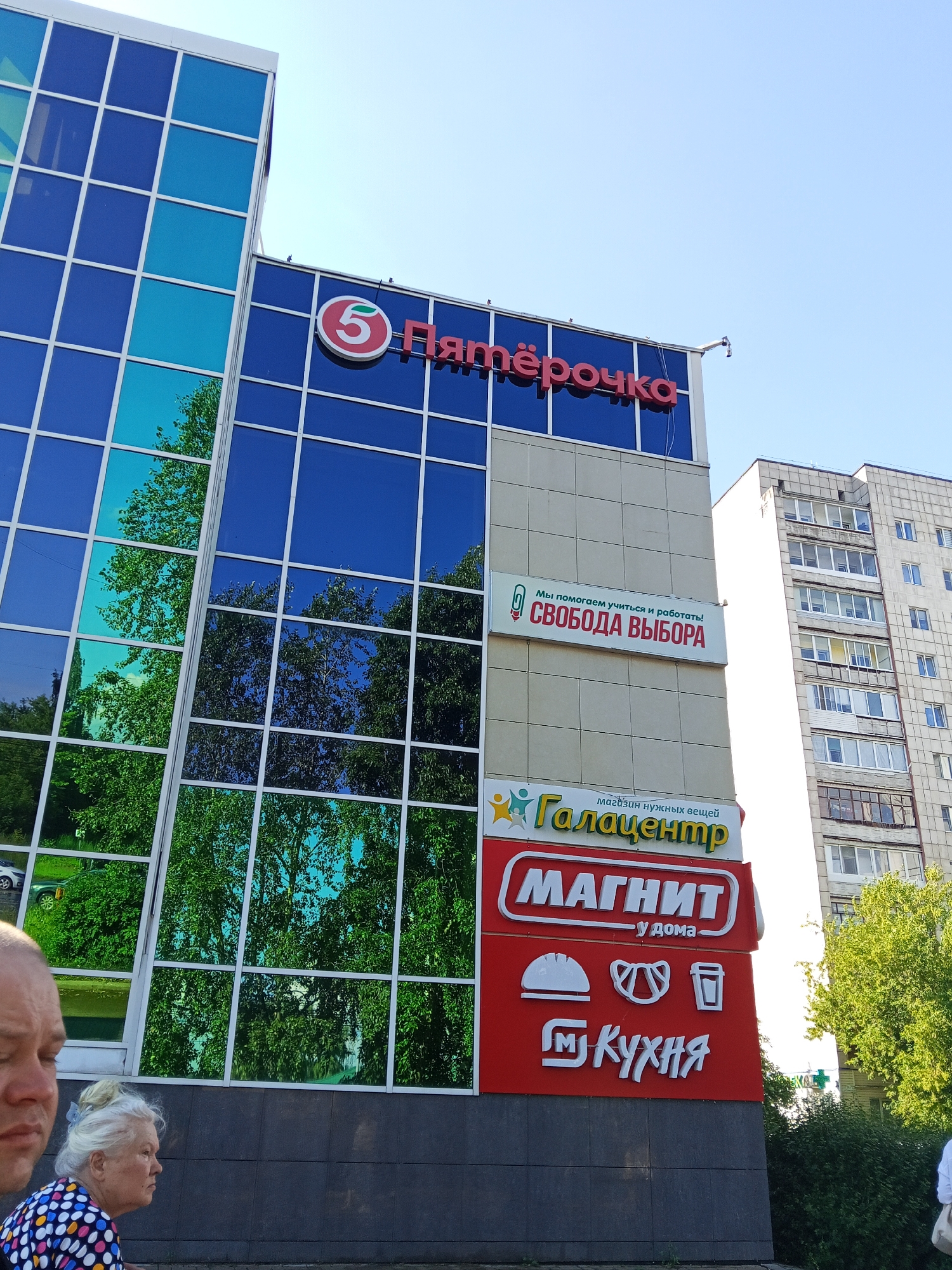 The image shows the exterior of a multi-story building with a blue glass facade. Several signs are visible on the building. One sign, with red lettering, reads "Пятёрочка" (Pyaterochka). Below this, another sign displays the words "Мы помогаем учиться и работать! СВОБОДА ВЫБОРА" (We help to study and work! Freedom of choice). Further down, a sign reads "Галацентр" (Galacenter), with smaller text below stating "магазин нужных вещей" (store of necessary things). The bottom sign is red with white lettering and logos, featuring "МАГНИТ" (Magnit), "у дома" (at home), and "КУХНЯ" (Kitchen). In the foreground, two individuals are partially visible: a man on the left and an older woman on the right, who is wearing a colorful polka-dot shirt. The background shows a tall apartment building. The scene appears to be a commercial area in the city of Chaikovsky, Russia.