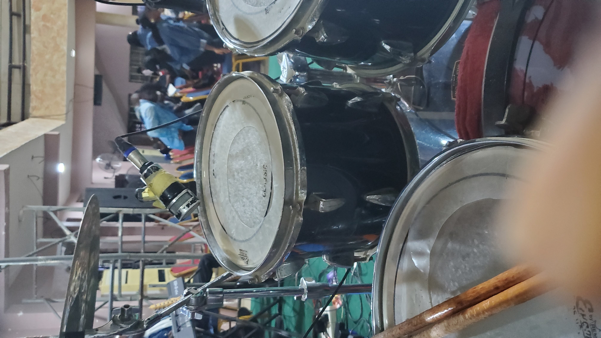 This is a medium shot photograph of a drum set in what appears to be an indoor event space in Jalingo, Nigeria. The foreground features two drumsticks resting on the rim of a drum. In the mid-ground, a black drum with a white resonant head is visible, with a microphone attached to its rim via a clamp. The microphone is wrapped in yellow tape. Behind the drum set, a crowd of people is visible, some wearing blue clothing. The background shows scaffolding, chairs, and a fan. The overall lighting suggests an indoor performance or gathering.