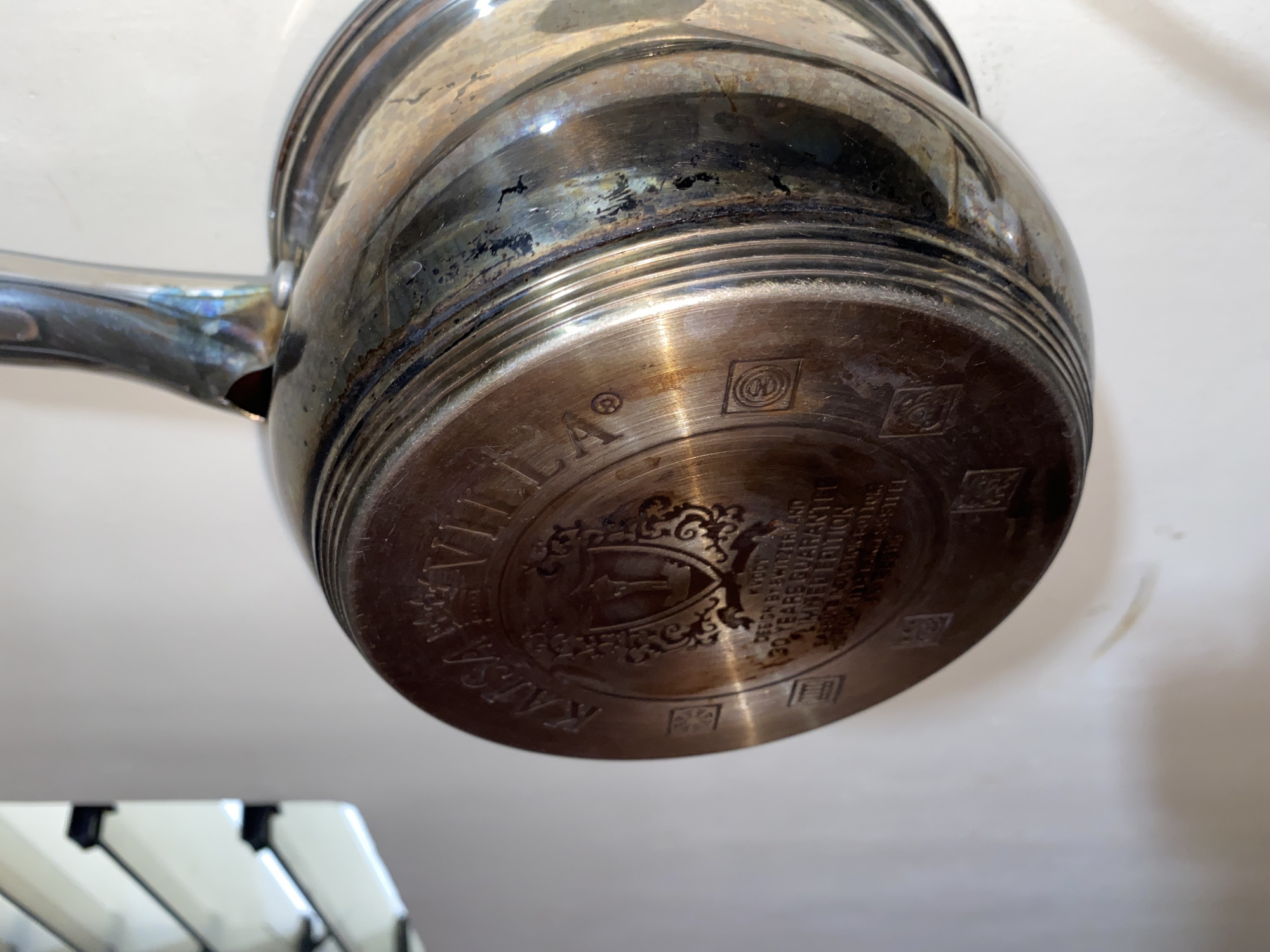 The image shows the bottom of a polished metal pot, presumably silver, held up to the light. The base of the pot is engraved with the name "KAISAVILLA," a crest or coat of arms with a hammer symbol, and the text "DESIGN BY SWITZERLAND 30 YEARS GUARANTY LIMITED EDITION." The surrounding area has small geometric decorative patterns. The pot appears to be old and shows signs of tarnishing. In the lower-left corner of the image, there is a partial view of what appears to be a white piano. The background is out of focus, but appears to be a white or light-colored wall. The image does not depict any location in Davao City, Philippines.