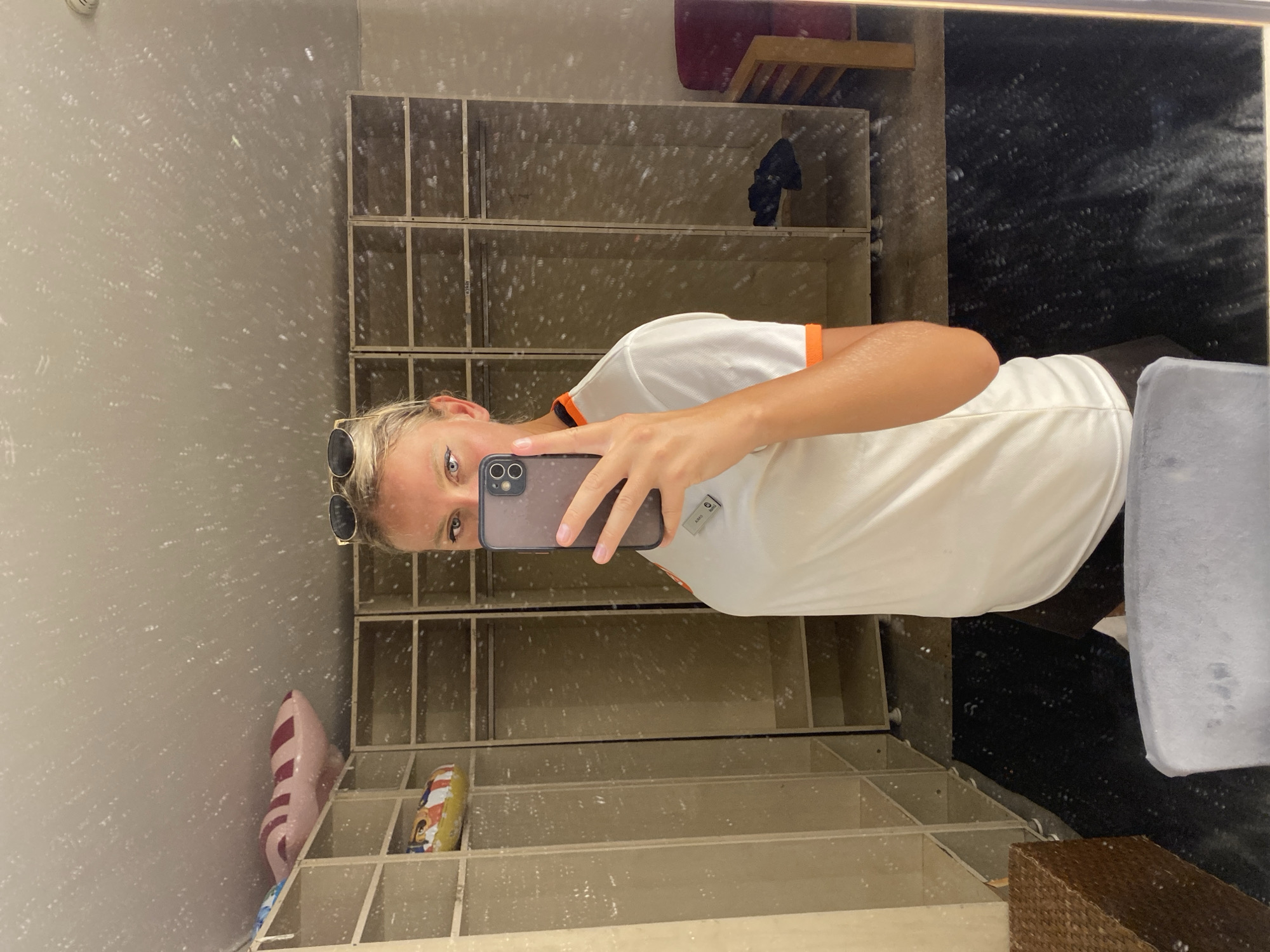 A woman takes a mirror selfie in what appears to be a locker room or dressing area, located in the city of Aksu, Turkey. She is wearing a white t-shirt with orange trim and dark shorts. She is holding a smartphone in her right hand and has sunglasses perched on her head. The background of the image reveals rows of empty lockers or storage cubbies. There are a few inflatable items on shelves in the upper left portion of the frame. A dark rug covers the floor in front of the lockers, and a wicker basket and a portion of a chair are visible in the foreground. The mirror's surface is visibly dirty, with numerous spots and smudges.