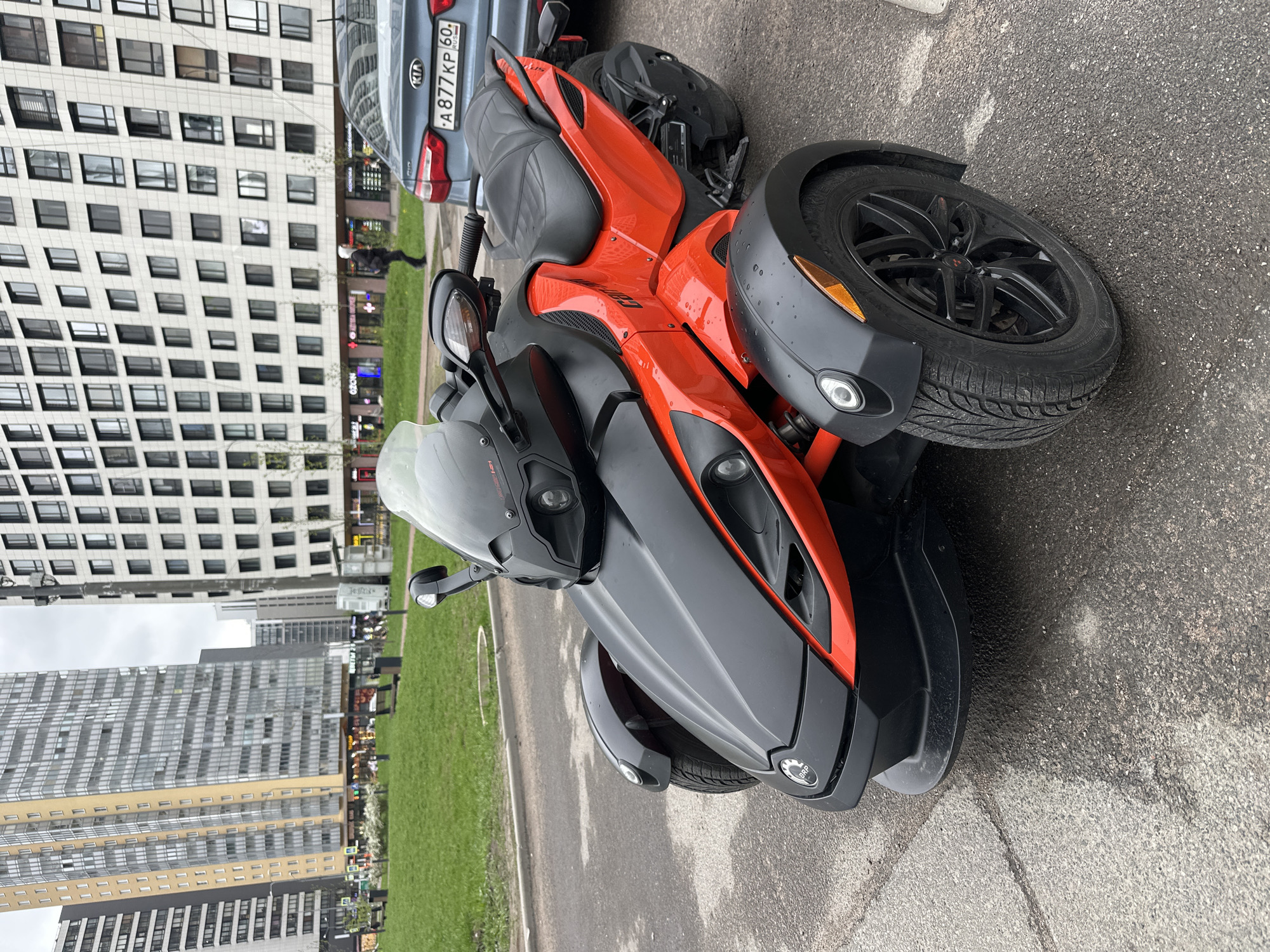 The image shows a Can-Am Spyder parked on asphalt. A gray car, a Kia, is partially visible behind it, with license plate A877 KP 60 RUS. In the background, there are modern apartment buildings, a green lawn, and some commercial storefronts at street level. Based on the architectural style and environment, the location is likely Murino, Russia. No people are clearly identifiable. The lighting suggests an overcast day.