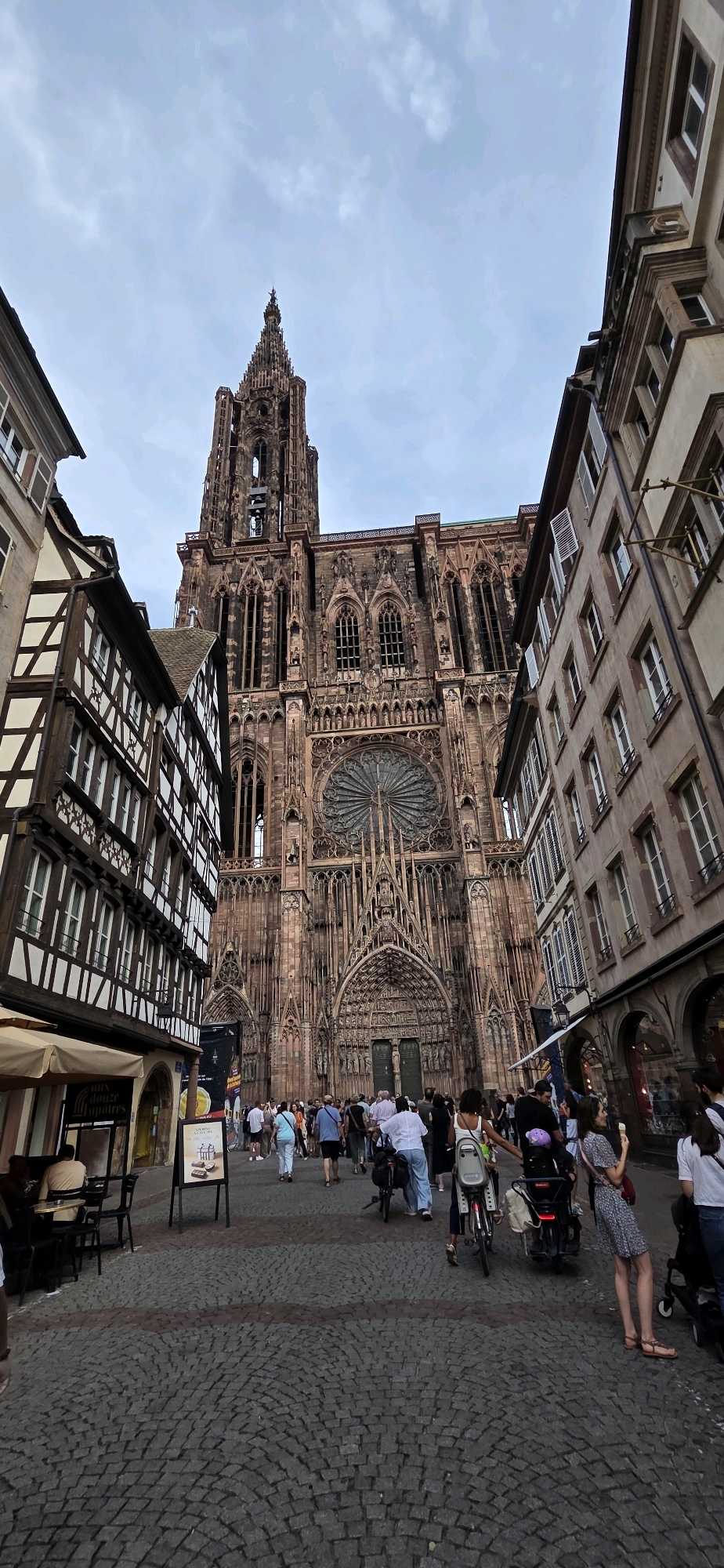 The image depicts a street scene in Strasbourg, France, with the Strasbourg Cathedral as the prominent backdrop. The cathedral, constructed of reddish stone, features a tall, intricate spire and a large rose window above its main entrance, which is adorned with numerous sculptures and gothic architectural details. Flanking the cathedral are buildings with traditional Alsatian architecture, characterized by their half-timbered facades and dark wooden beams against lighter plaster.
A cobblestone street with a patterned walkway occupies the foreground and middle ground. A crowd of people are present in the scene, walking, standing, and interacting. Some individuals are pushing bicycles or strollers, suggesting a public space that is actively used by pedestrians. Several people are dressed in casual, summery attire. There are also outdoor seating areas for cafes or restaurants visible to the left, with patrons seated at tables. A street sign with text and an image is also present. The sky is overcast, indicating a cloudy day. The overall atmosphere is that of a busy, historic urban environment.