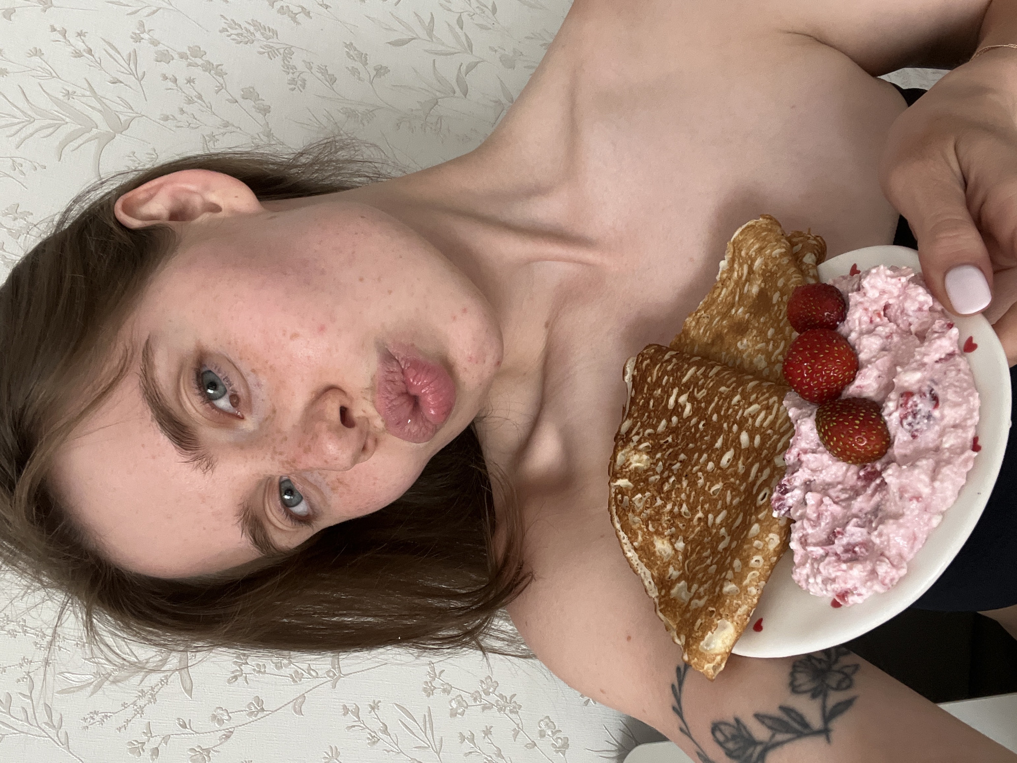The media file shows a close-up, overhead shot of a person with fair skin and freckles, lying down with their head tilted. They have light blue eyes and are looking towards the camera with their lips puckered. Their dark brown hair is spread around their head. To the right of the person's face is a white plate containing two thin pancakes and a portion of pink cottage cheese with visible pieces of fruit, likely strawberries. Three whole strawberries are also on the plate, two on top of the cottage cheese. A tattoo of a floral branch is visible on the person's arm. The background is a light-colored wallpaper with a subtle floral pattern. The lighting suggests it is daytime, possibly indoors. The person appears relaxed, with a playful expression. No text is visible in the image.
