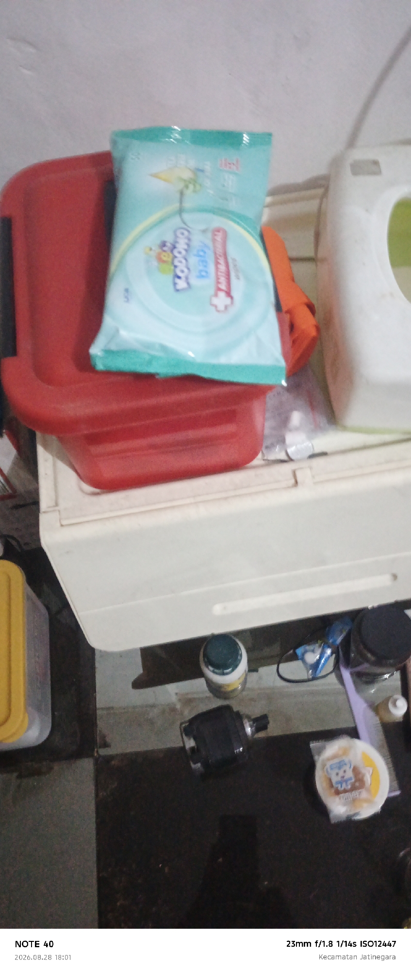 The image shows a close-up of several objects on a tiled surface, with a white plastic container and a red plastic container visible in the background. A package of baby wipes, identified by the text "MOMMY BABY" and a bear illustration, is placed on top of the red container. To the right of the baby wipes, an orange object is partially visible. Further in the background, a white appliance is present. On the tiled surface in the foreground, a small white and green capped bottle, a black electronic device, and a circular package with a cartoon bear design are discernible. The bottom of the image contains a timestamp "2026.08.28 18:01" and a location "Kecamatan Jatinegara," indicating East Jakarta, Indonesia.