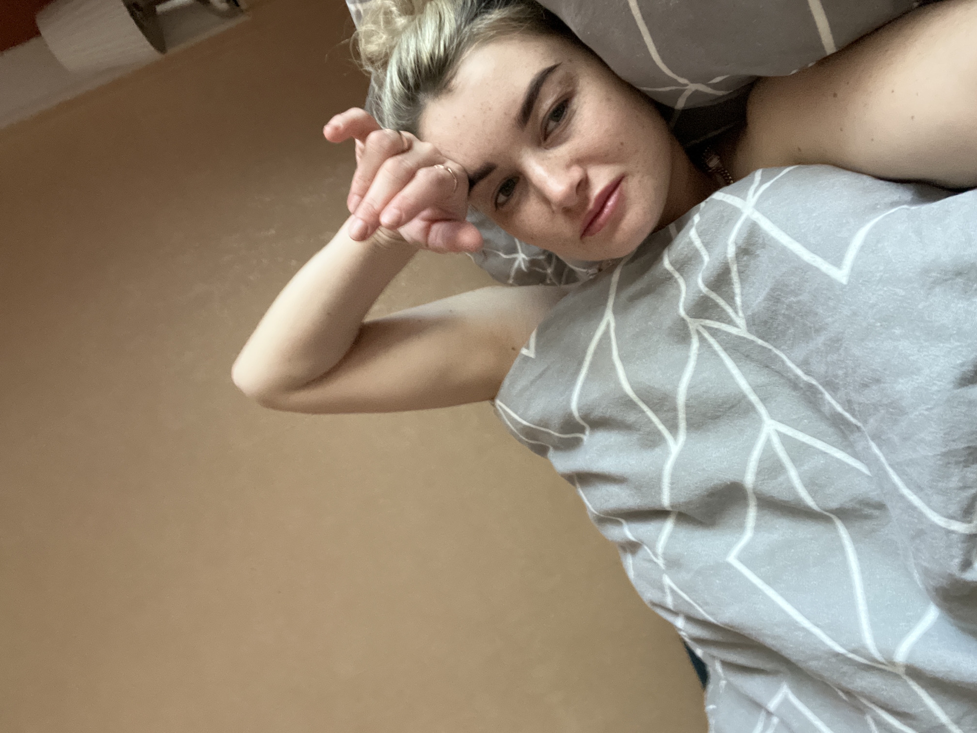 A woman is lying in bed, appearing tired or waking up. Her blond hair is pulled up in a messy bun. She is partially covered by a gray comforter patterned with white geometric shapes, and a gray pillow is behind her head. She has one hand resting on her forehead. The background shows a tan-colored wall and a corner of a white object (possibly a fixture or outlet). No specific location in Chaikovsky, Russia is indicated.