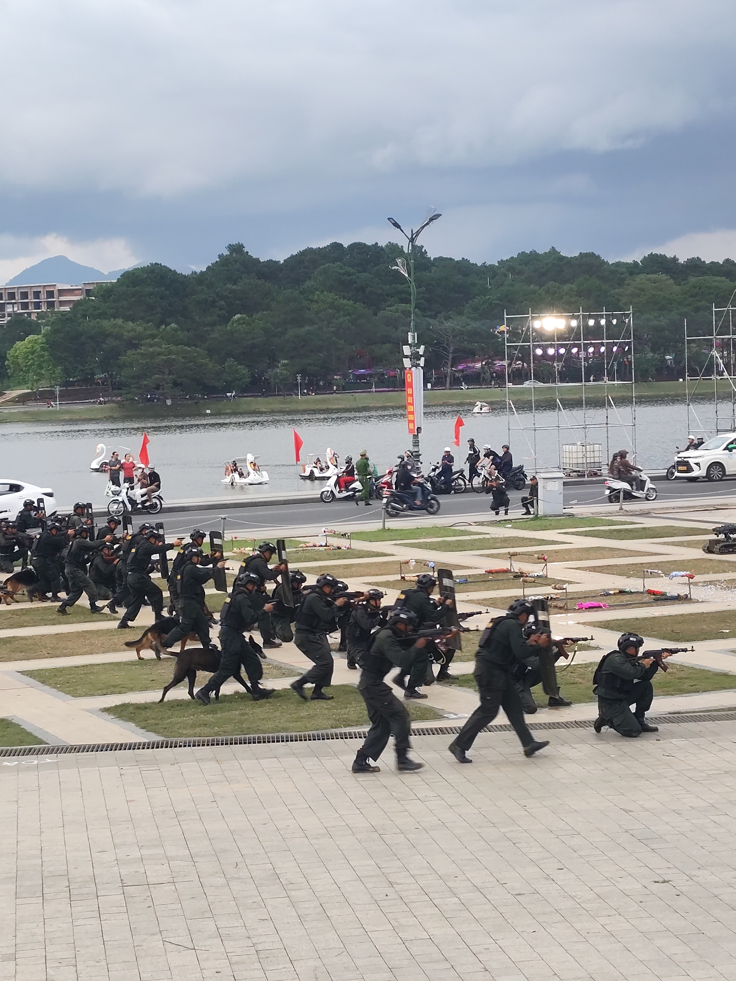 In Da Lat, Vietnam, a scene unfolds near Xuan Huong Lake. A group of individuals, dressed in tactical gear including helmets, vests, and dark uniforms, are engaged in what appears to be a training exercise or demonstration. They are armed with rifles and shields and are depicted in various dynamic poses, suggesting movement and action, such as running and aiming. Several German Shepherd dogs, also in tactical harnesses, are present and appear to be moving with the group, likely trained for security or apprehension.
The environment includes a paved walkway in the foreground, with a grassy area beyond. In the mid-ground, the calm waters of Xuan Huong Lake are visible, with several swan-shaped pedal boats and a few people on them. On the opposite side of the lake, a backdrop of lush green trees and hills can be seen, with some buildings visible on the slopes. A stage with lighting equipment is set up near the water's edge, suggesting a potential event or performance. A white sedan and several motorcycles are also present near the lake, indicating vehicular traffic. The sky is overcast, with a hint of dramatic clouds, suggesting a day with variable weather. The overall impression is a public demonstration or training exercise involving law enforcement or military personnel in a scenic lakeside setting.