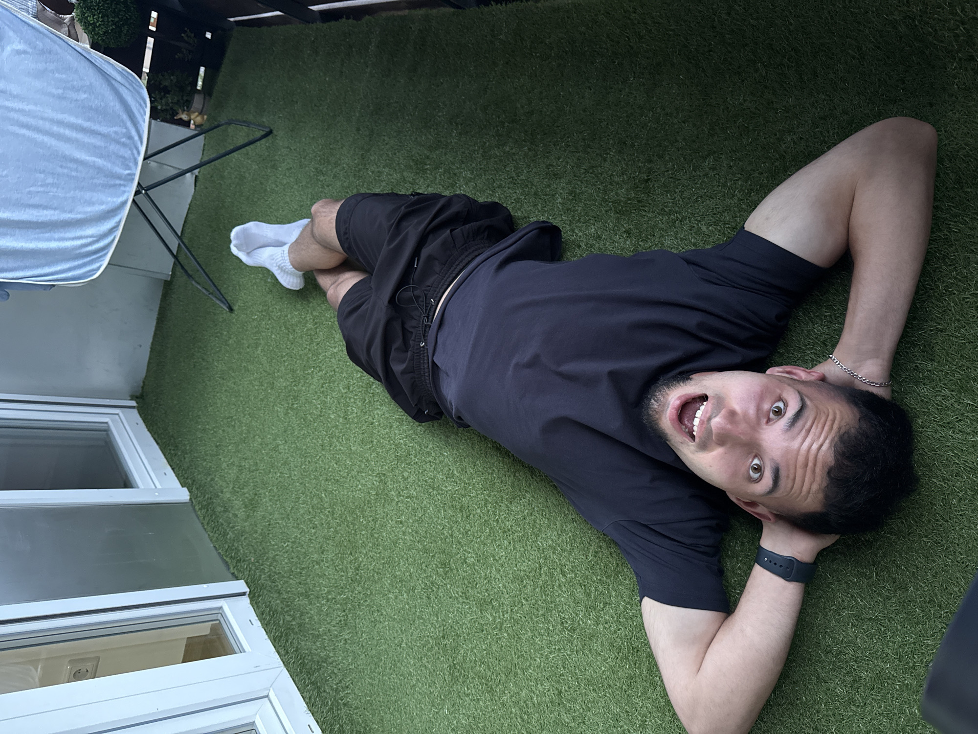 A male individual is lying supine on green artificial turf on a balcony or patio in Garmisch-Partenkirchen, Germany. He is dressed in a dark short-sleeved t-shirt and dark shorts, with white athletic socks. His hands are clasped behind his head, and he looks directly towards the camera with wide eyes and an open mouth, conveying a surprised or playful expression. A dark smartwatch is visible on his left wrist and a silver chain bracelet on his right. In the background to the left, white-framed glass doors or windows lead to an interior space, revealing a power outlet on the interior wall. To the right, a light blue fabric sheet is draped over a black metal clothes drying rack. Further right, a small potted plant and a decorative figurine are visible. The scene depicts a relaxed moment in an outdoor residential setting.