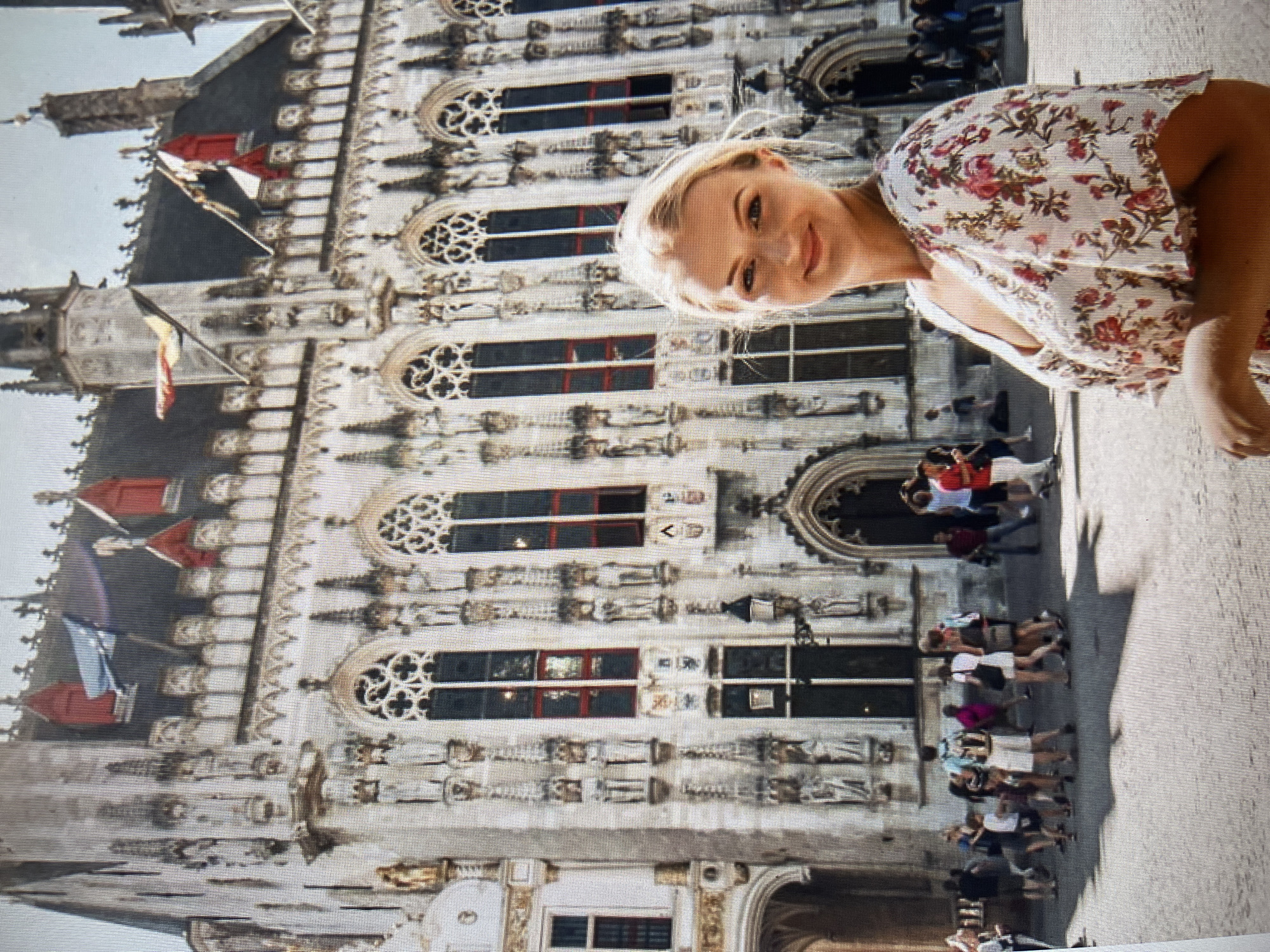 The image shows a woman in a floral dress standing in front of a large, ornate building. The building appears to be a historical landmark with intricate stonework and multiple windows. Several flags are visible on the roof of the building. In the background, there are other people walking around and a cobblestone plaza. The woman is smiling and looking towards the camera. The lighting suggests it is daytime, possibly a sunny day. The scene depicts a tourist taking a photograph in front of a notable architectural structure. The location is not Qarağandı city administration, Kazakhstan.